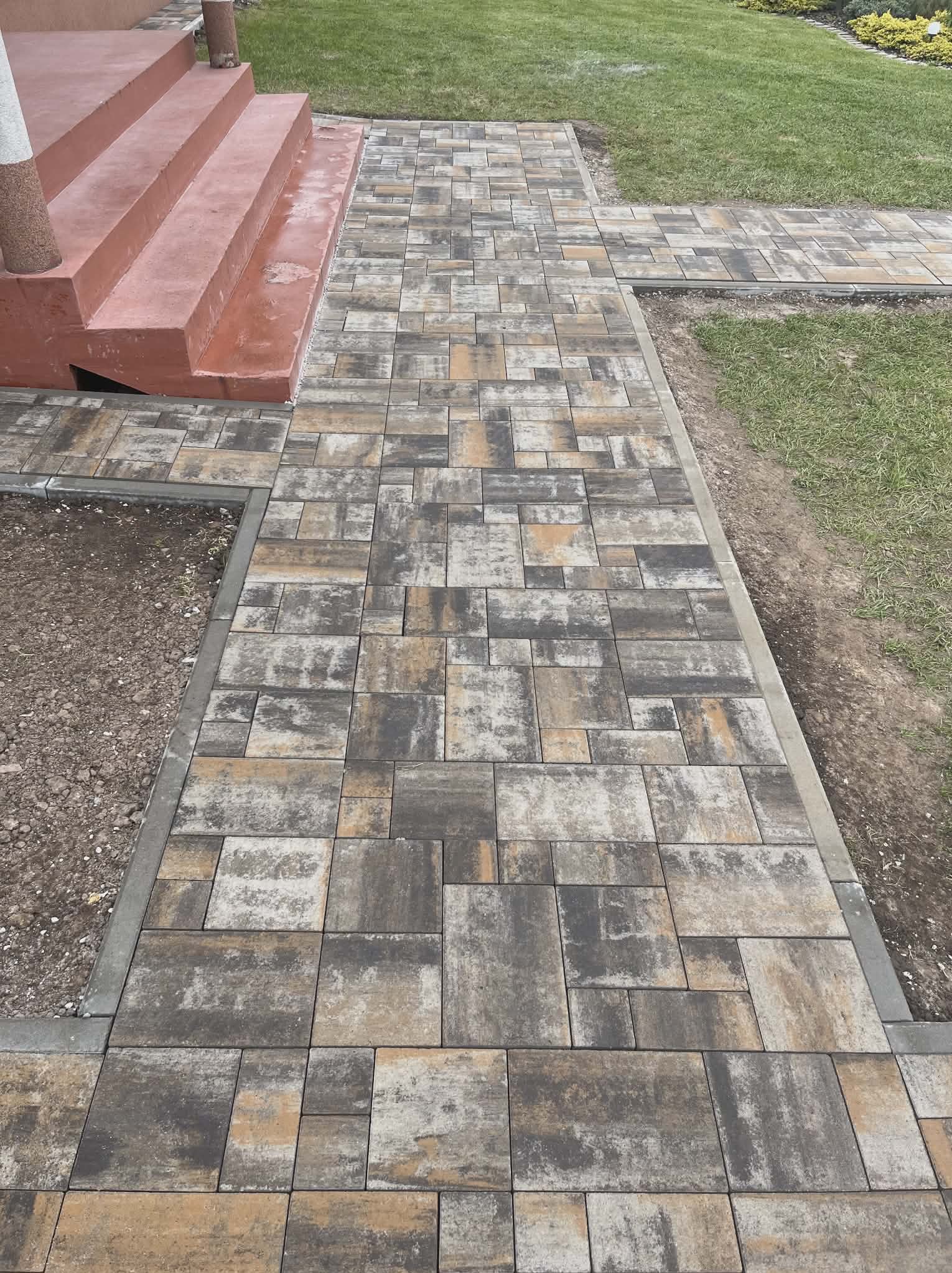 Ścieżka z kostki brukowej w odcieniach szarości i beżu, prowadząca od schodów do trawnika. Krawężniki betonowe. Widok z góry, lekko z ukosa.