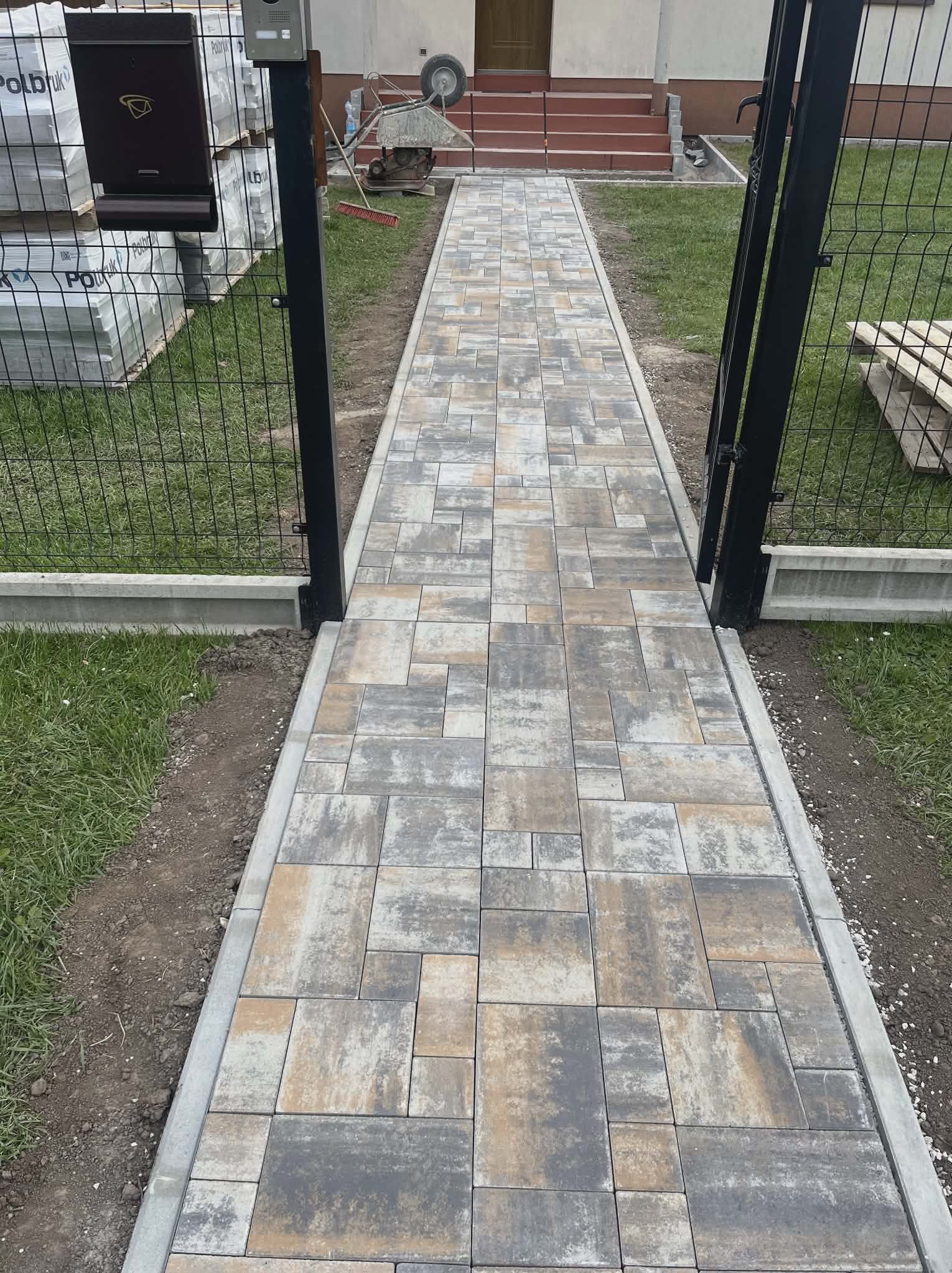 Ułożona ścieżka z kostki brukowej prowadząca przez bramkę ogrodzeniową, z widocznymi paletami kostki w tle. Różnokolorowa kostka tworzy wzór na ścieżce.