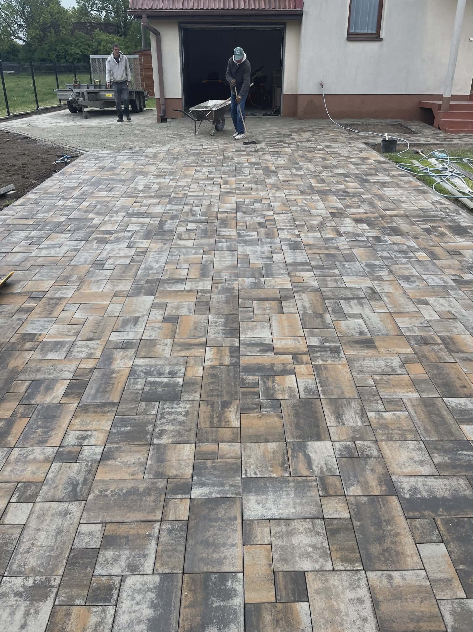 Ułożona kostka brukowa w odcieniach szarości i beżu na podjeździe przed domem. Widoczni pracownicy i narzędzia. Perspektywa z dołu podkreśla wzór kostki.