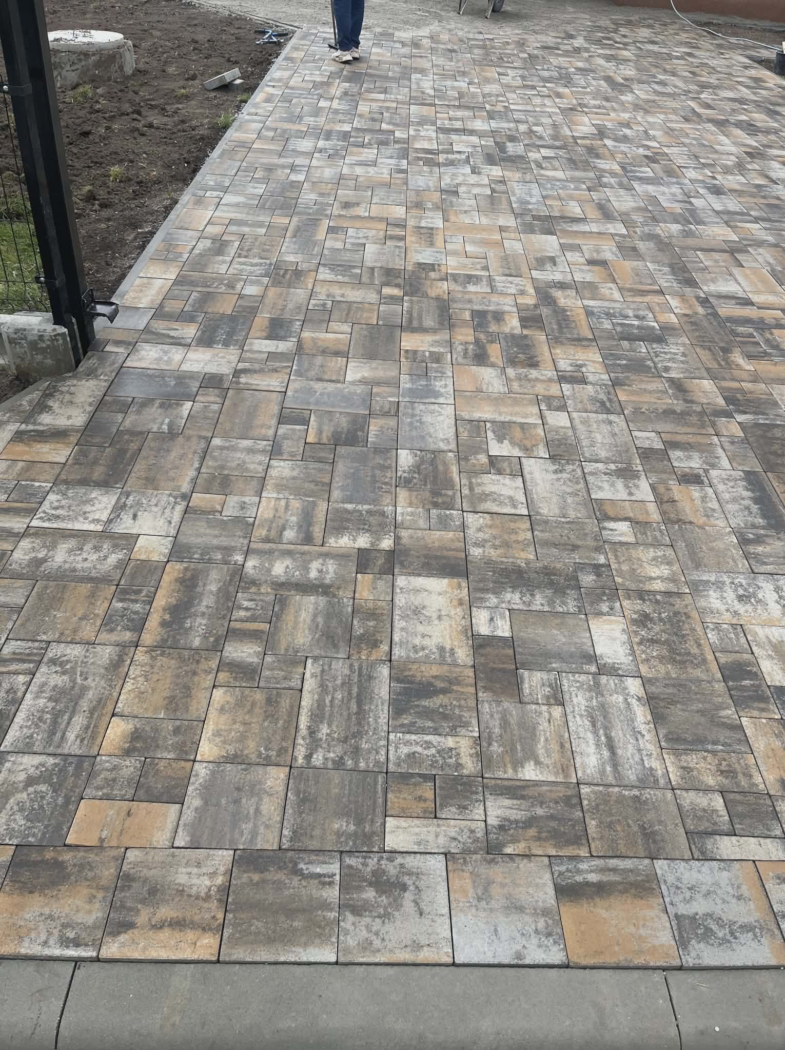 Ułożona kostka brukowa w odcieniach szarości i beżu, tworząca wzór na podjeździe. Widoczne obrzeża i fragment ogrodzenia. Perspektywa z góry.