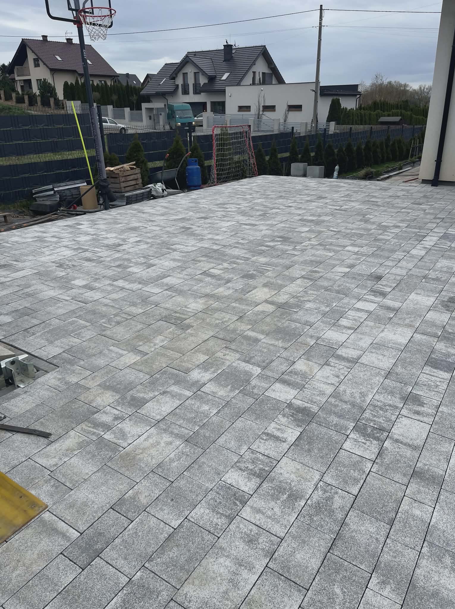 Ułożona szara kostka brukowa na posesji, widok z góry. W tle domy, ogrodzenie i elementy wyposażenia ogrodu: bramka do piłki nożnej i kosz do koszykówki.