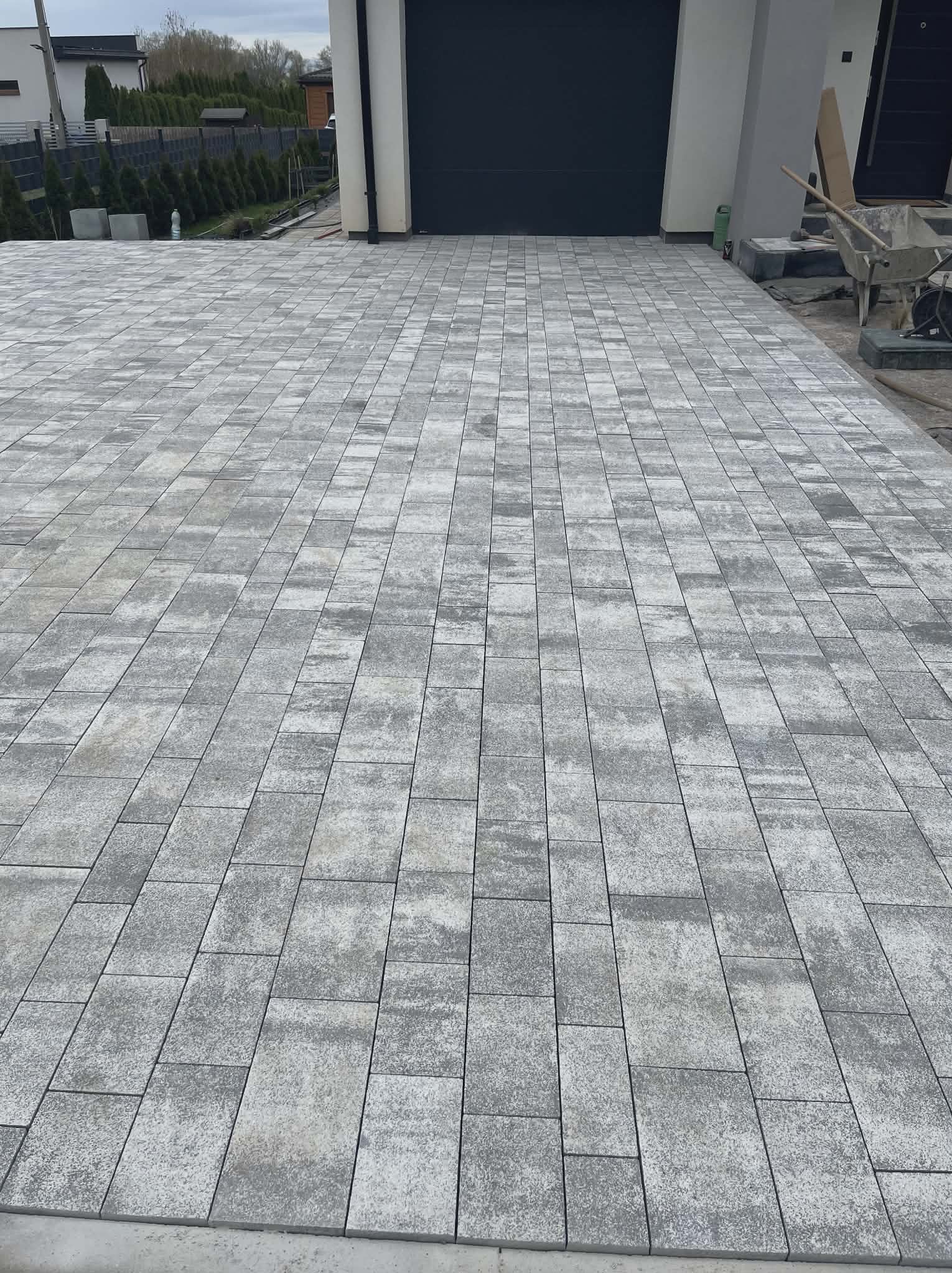 Ułożona, szara kostka brukowa na podjeździe przed garażem z czarną bramą. Widoczny fragment domu i roślinność w tle. Perspektywa z dołu, podkreślająca wzór kostki.
