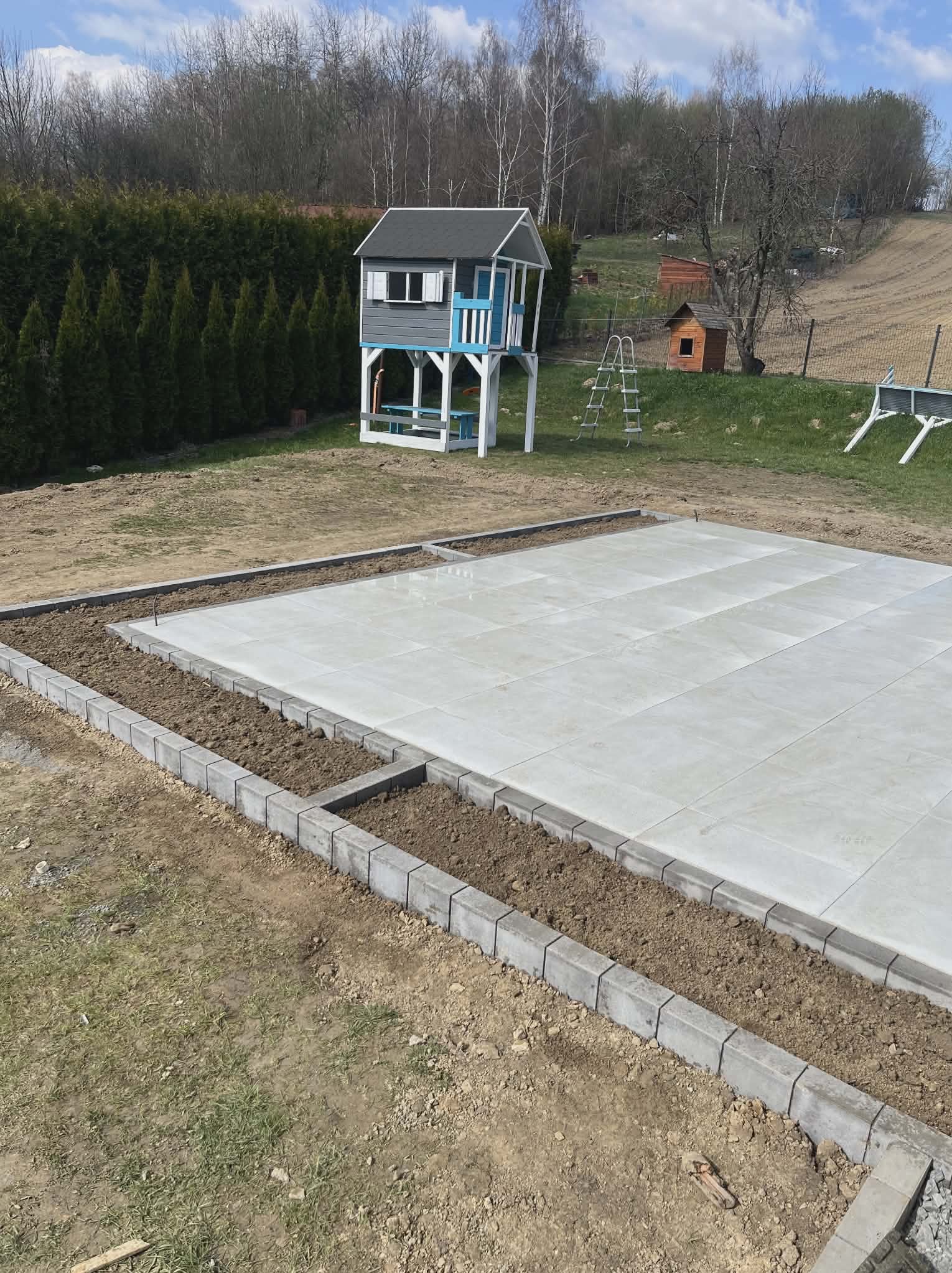 Jasnoszara, kwadratowa nawierzchnia z kostki, obrzeża z szarej kostki, ziemia. W tle domek dla dzieci na podwyższeniu i zieleń. Słoneczny dzień.