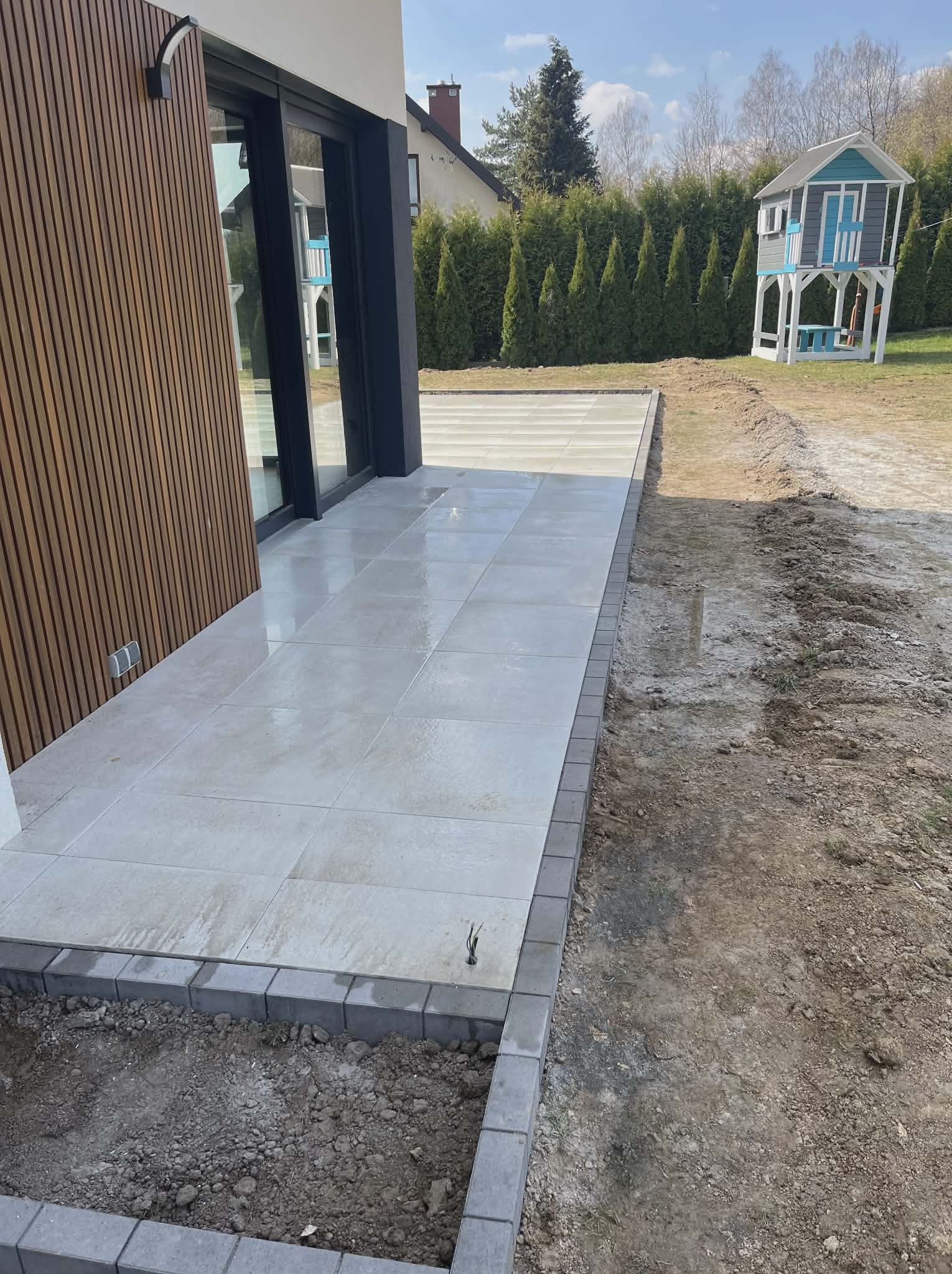 Jasnoszara, kwadratowa kostka brukowa ułożona przy nowoczesnym domu z drewnianą elewacją. W tle zielony trawnik i domek dla dzieci.