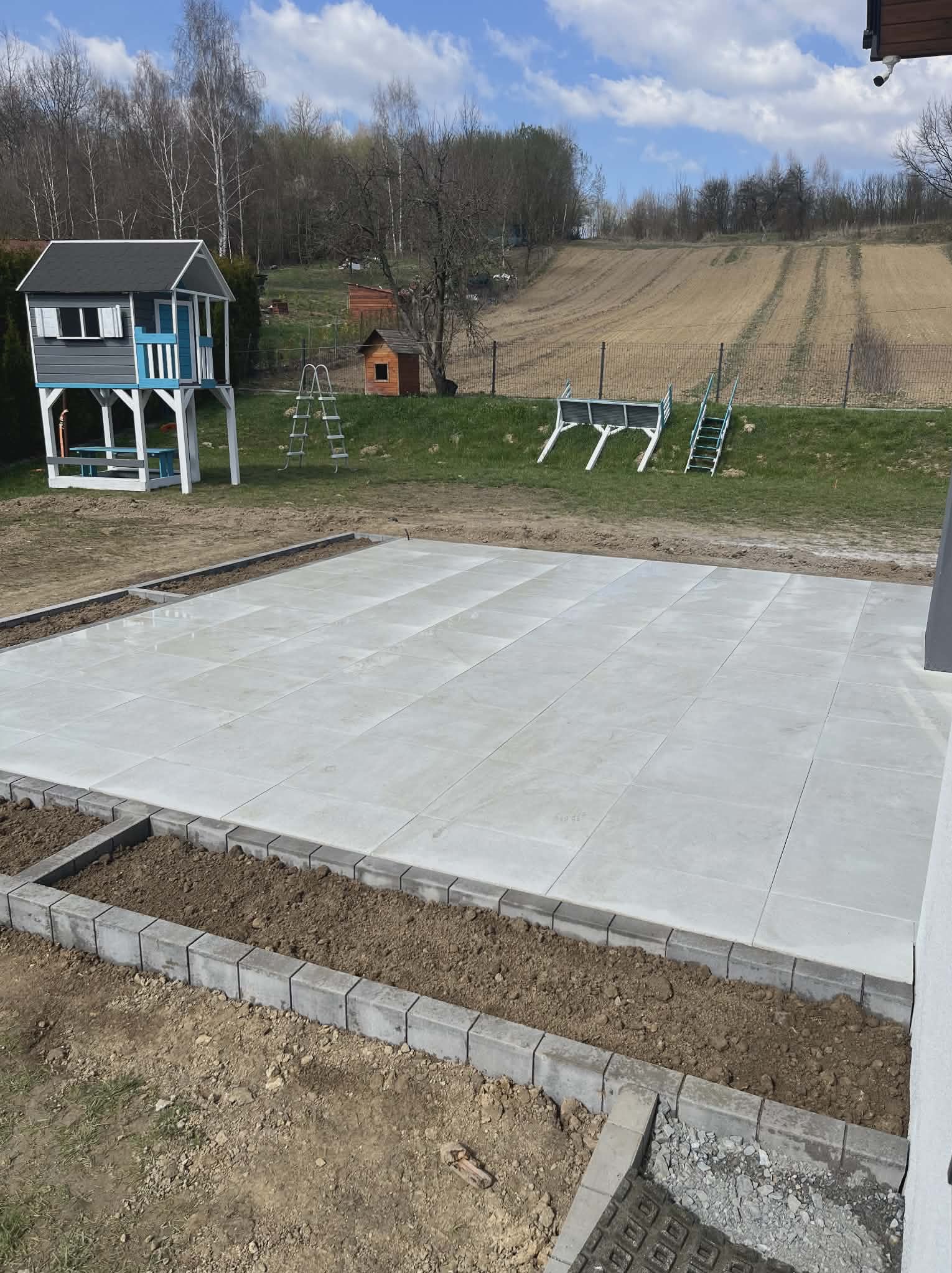 Jasnoszara, duża powierzchnia z płyt betonowych obramowana kostką, przygotowana pod nasadzenia. W tle plac zabaw i pole uprawne pod błękitnym niebem.