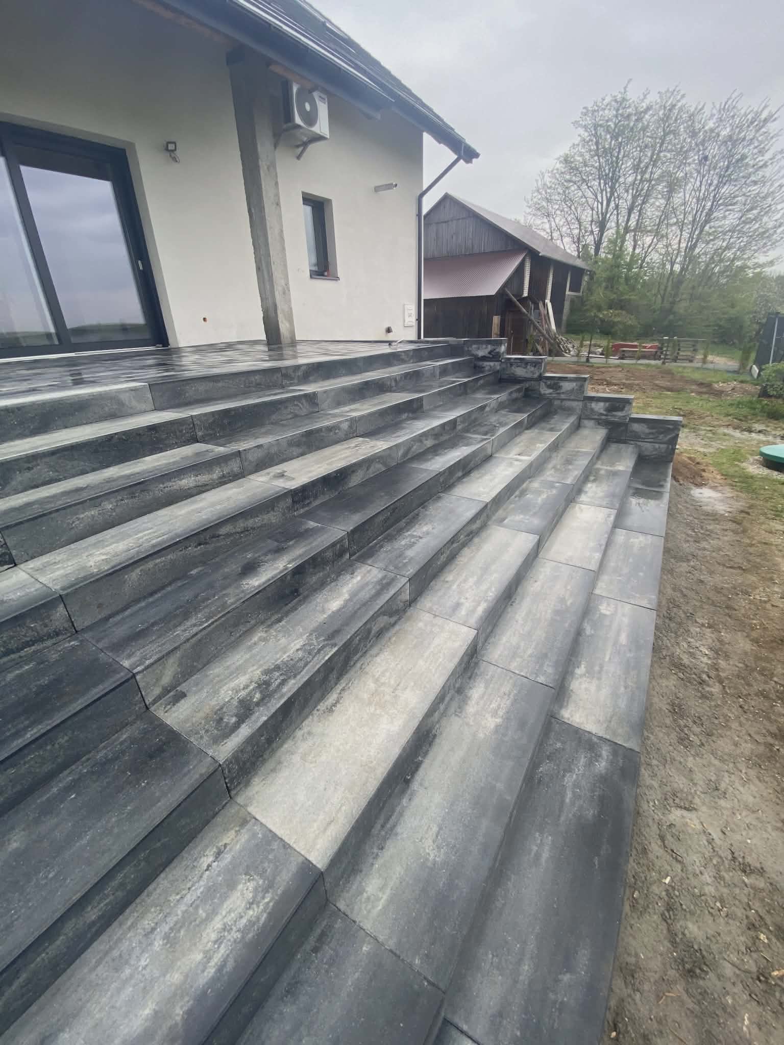 Schody z kostki brukowej prowadzące do domu z klimatyzatorem na ścianie, w tle budynek gospodarczy i drzewa. Kostka w odcieniach szarości, ułożona z precyzją.