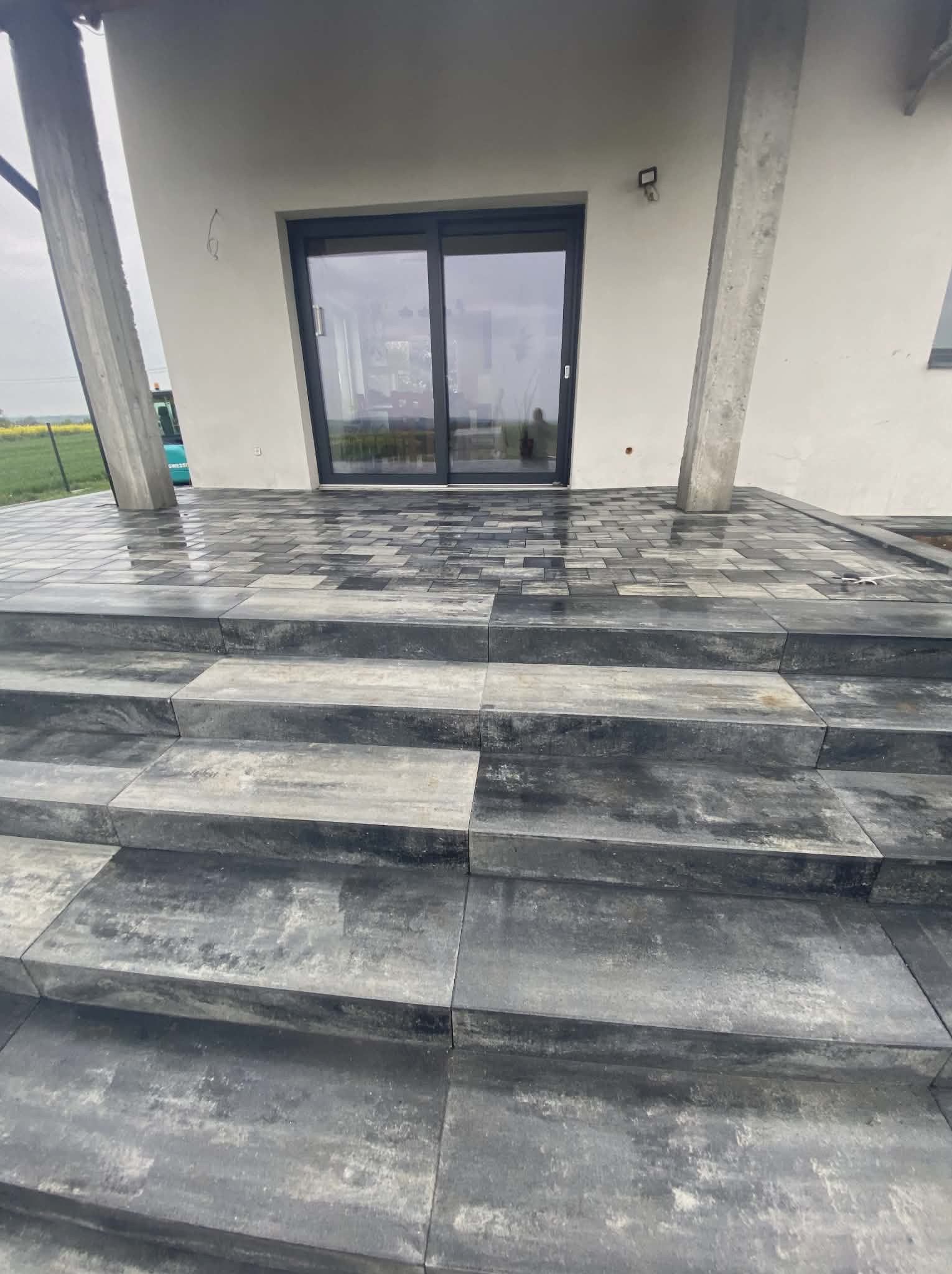 Nowoczesne schody wejściowe i taras z szarej kostki brukowej, widok z dołu, zarys budynku w tle, pochmurne niebo. Kostka ułożona w geometryczny wzór.