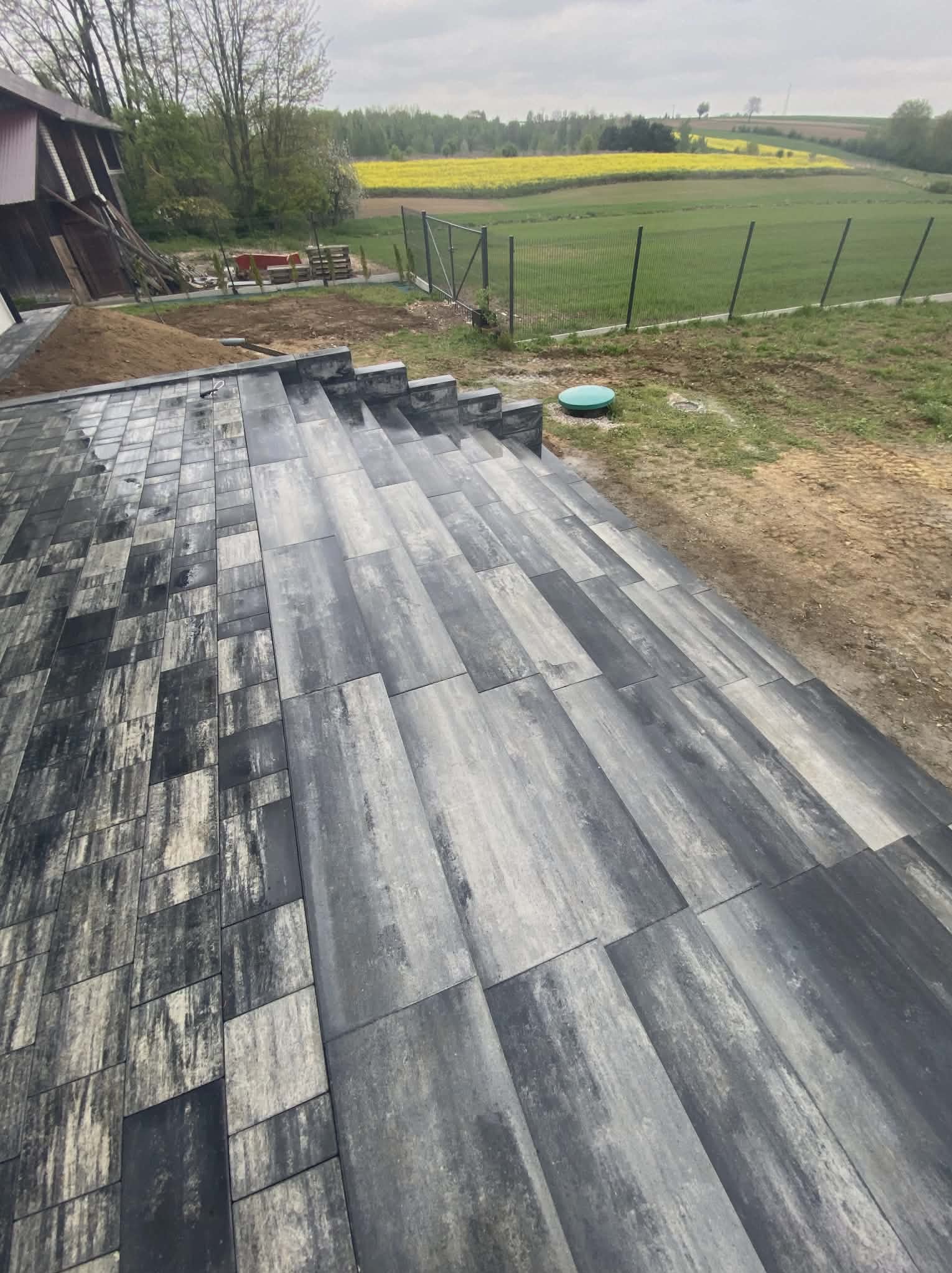 Ułożona kostka brukowa w Iwanowicach Włościańskich, tworząca schody na nierównym terenie. Widok na pola i łąki w tle. Kostka w odcieniach szarości, ułożona w regularny wzór.