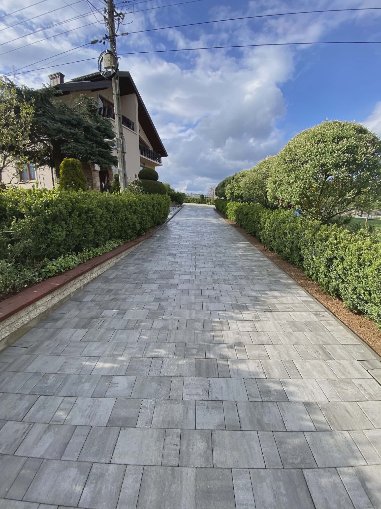 Długa, szara ścieżka z kostki brukowej prowadząca do domu, otoczona zielenią i krzewami. Widok perspektywiczny, niebo z chmurami w tle.