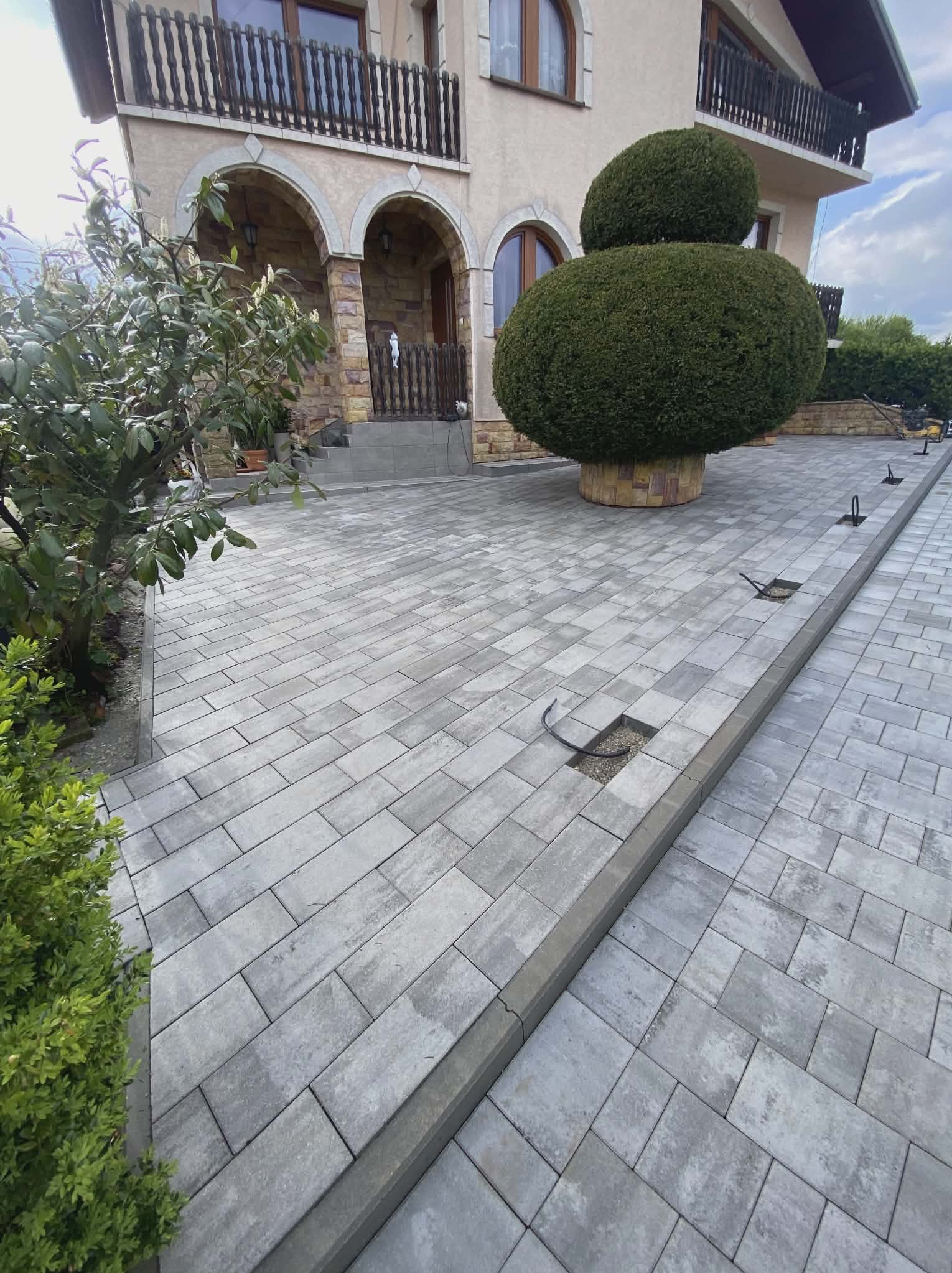 Ułożona szara kostka brukowa na podjeździe przed domem z ozdobnym krzewem i balkonem. Widoczne niedokończone łączenia kostki.