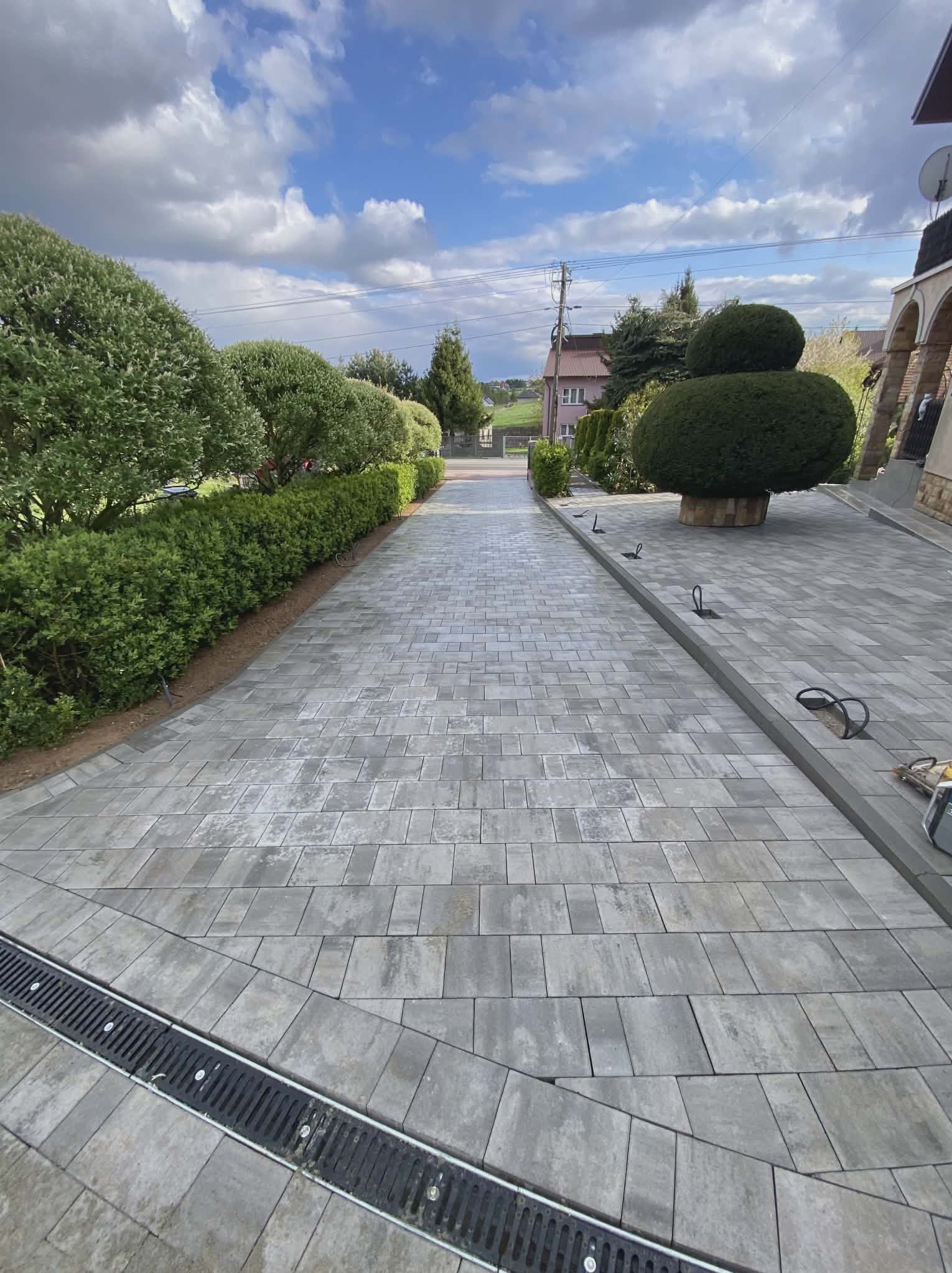 Ułożona szara kostka brukowa tworząca ścieżkę w ogrodzie, z widocznym odpływem liniowym i starannie przyciętymi krzewami po bokach. Słoneczny dzień.