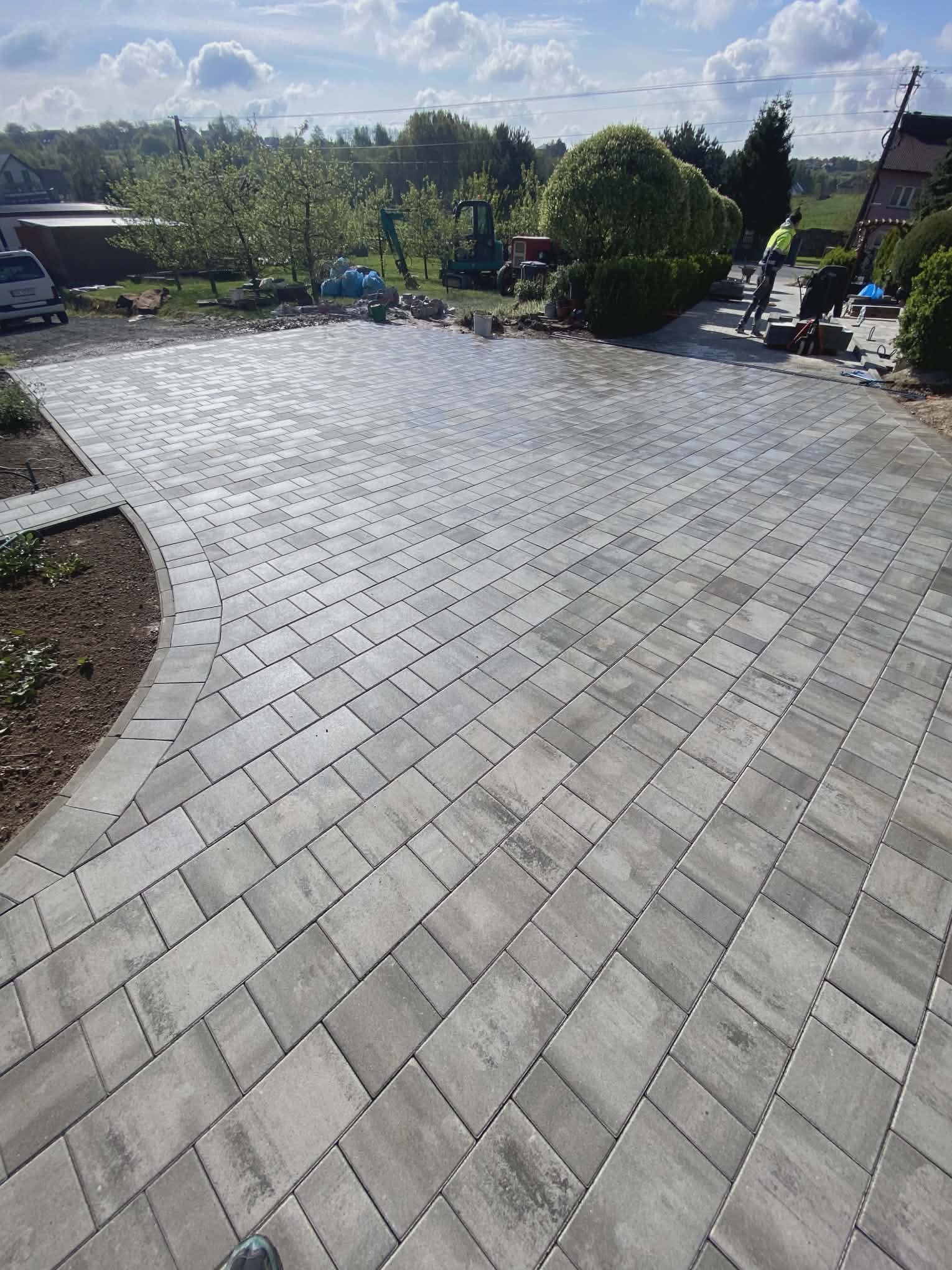 Ułożona szara kostka brukowa na podjeździe, widoczny fragment ogrodu i maszyny budowlane w tle, częściowo widoczna stopa osoby w lewym dolnym rogu.
