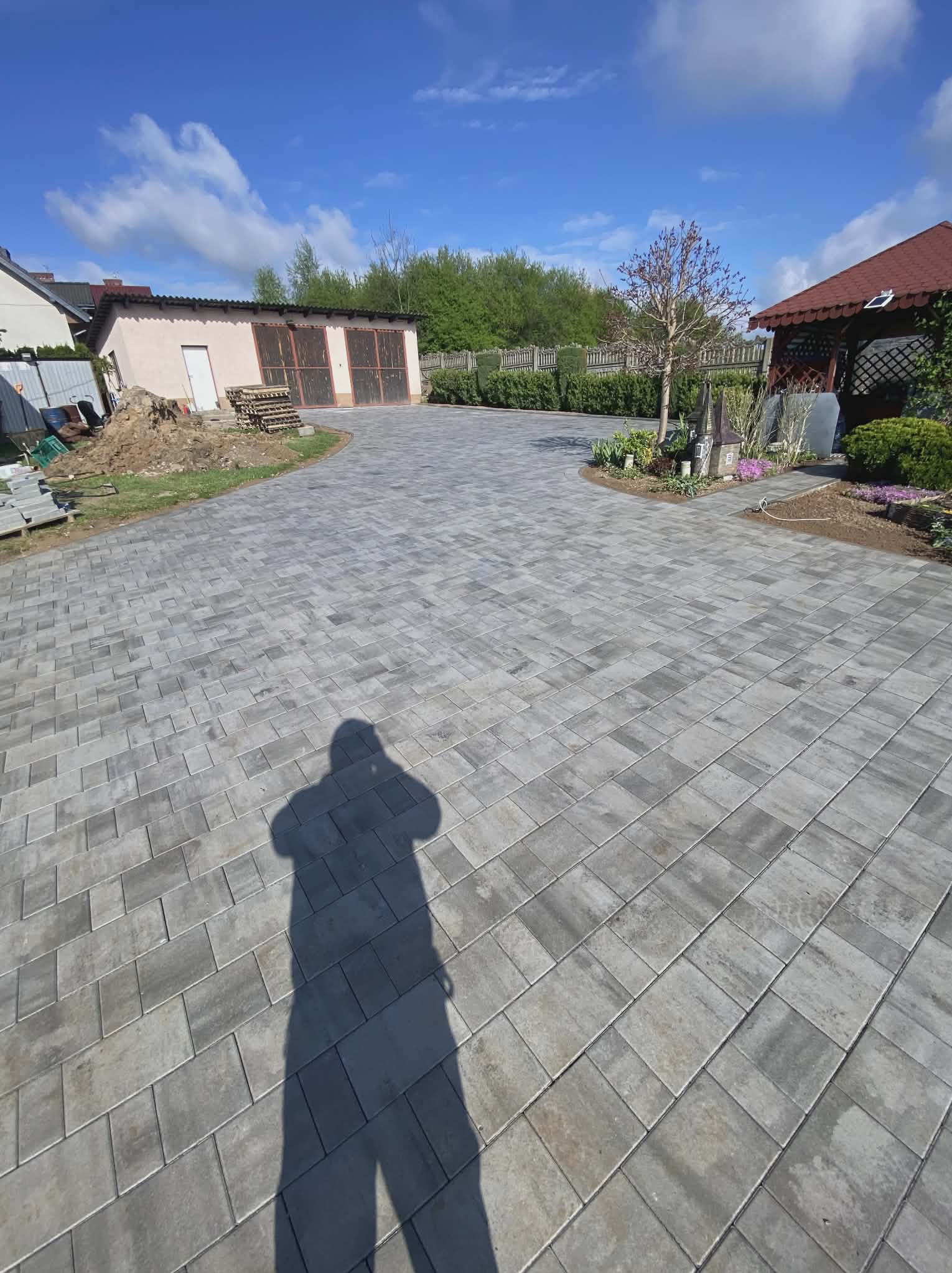Szara kostka brukowa ułożona na podjeździe, cień osoby w kadrze, w tle budynek gospodarczy i altana. Widok na posesję w słoneczny dzień.