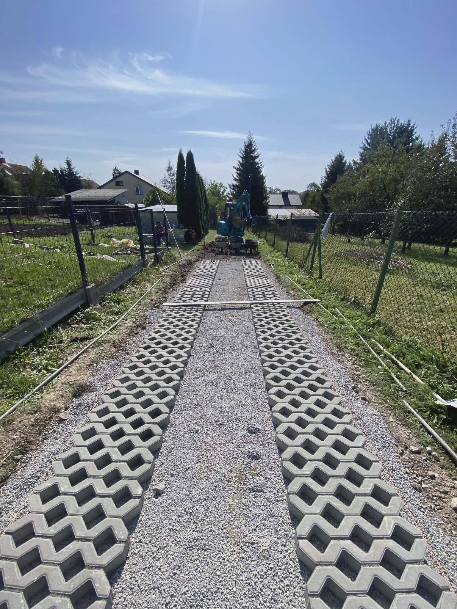 Układanie ażurowej kostki brukowej na podbudowie z kruszywa. W tle widoczny mały sprzęt budowlany i ogrodzenie posesji. Prace w toku, dzień słoneczny.