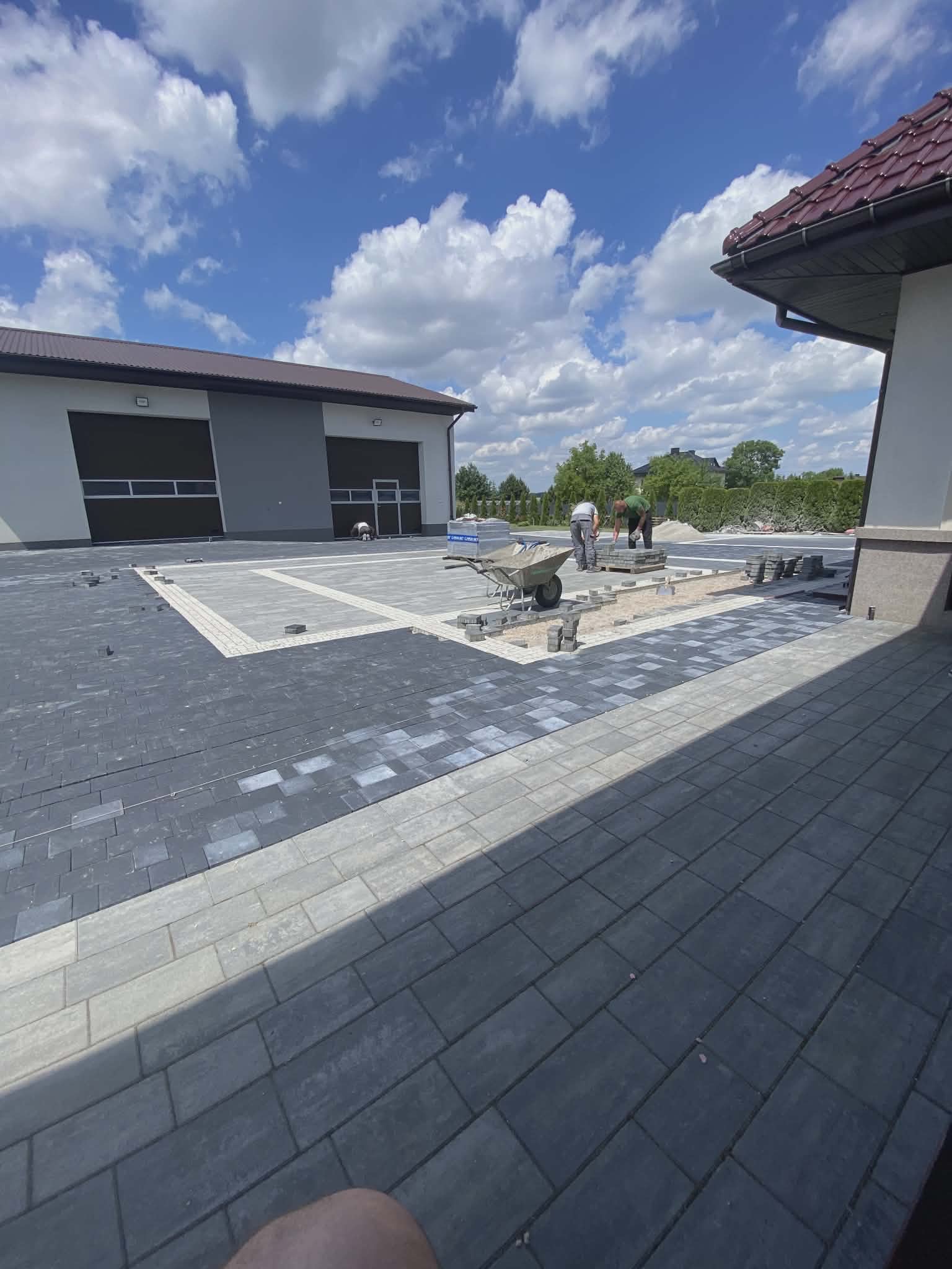 Układanie ciemnoszarej kostki brukowej z jasnymi obrzeżami na posesji. Widoczni pracownicy i taczka z materiałami. Częściowo ułożona nawierzchnia, błękitne niebo z chmurami.