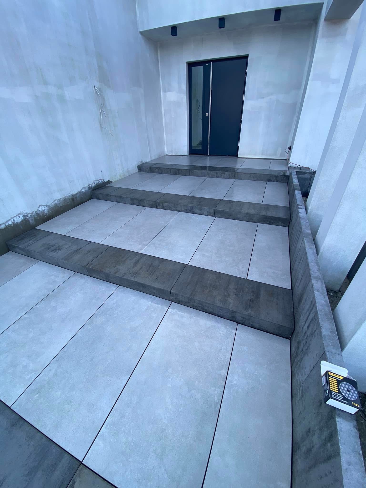 Nowoczesne schody wejściowe wyłożone jasnoszarą, prostokątną kostką brukową z ciemniejszymi obrzeżami, prowadzące do ciemnych drzwi wejściowych.