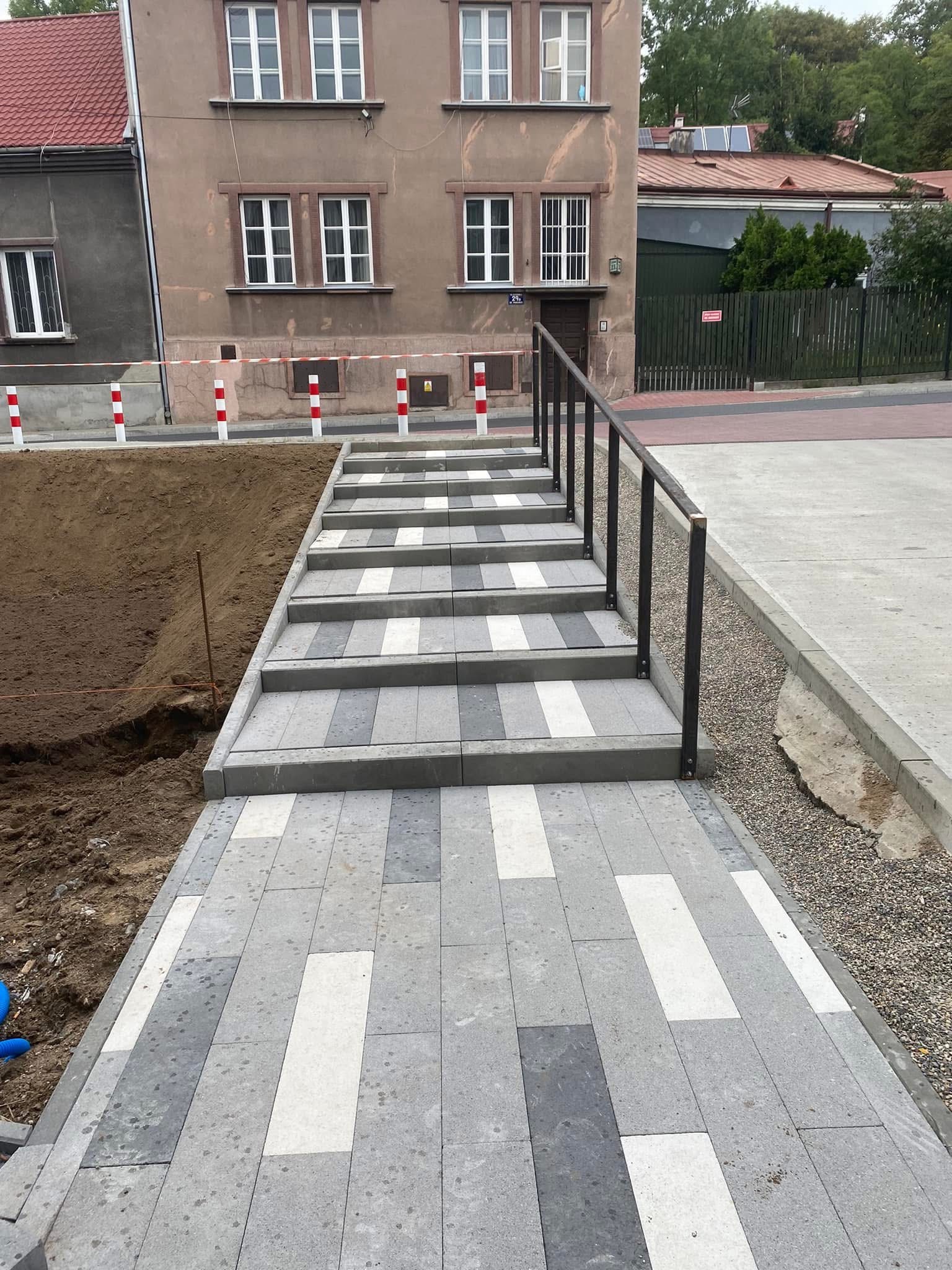 Schody z kostki brukowej w odcieniach szarości i bieli, prowadzące do budynku. Metalowa barierka po prawej stronie. Obok widoczny wykop ziemny.