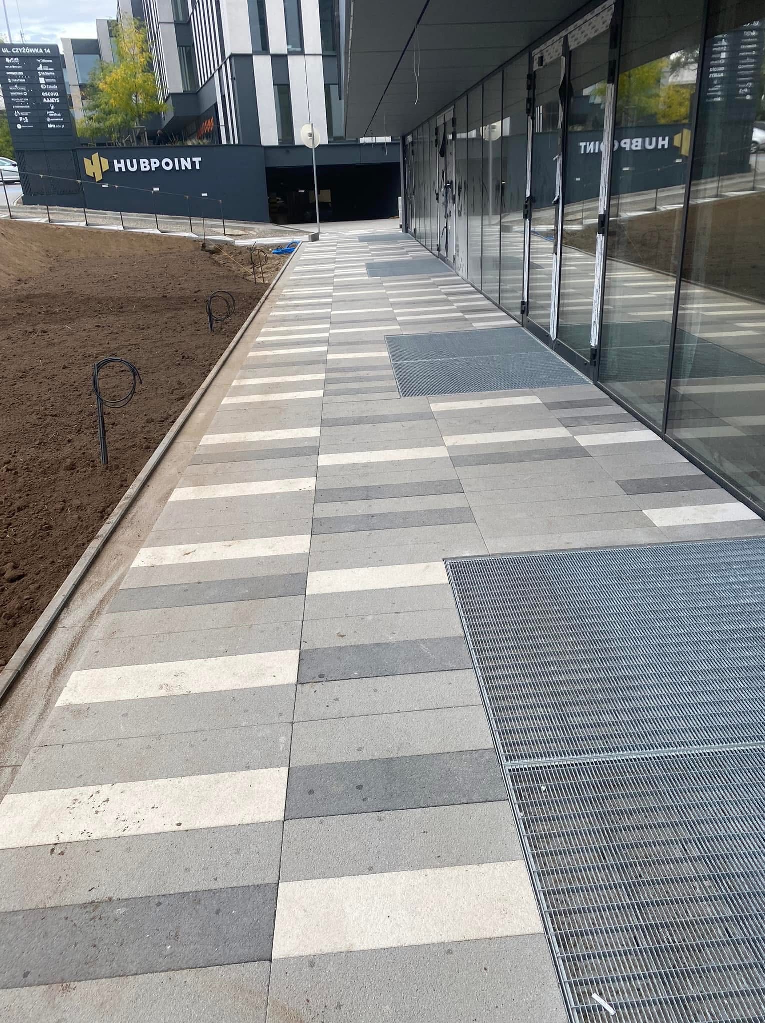 Nowoczesny chodnik z kostki brukowej w odcieniach szarości i bieli, z kratkami odpływowymi, przylegający do budynku biurowego z przeszkloną fasadą.