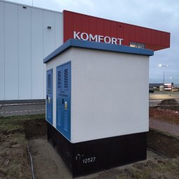 CREATO GROUP SPÓŁKA Z OGRANICZONĄ ODPOWIEDZIALNOŚCIĄ - Nowo postawiona, biało-czarna stacja transformatorowa z niebieskimi drzwiami wentylacyjnymi, umieszczona na placu budowy przed budynkiem sklepu Komfort, widoczne kable w ziemi.
