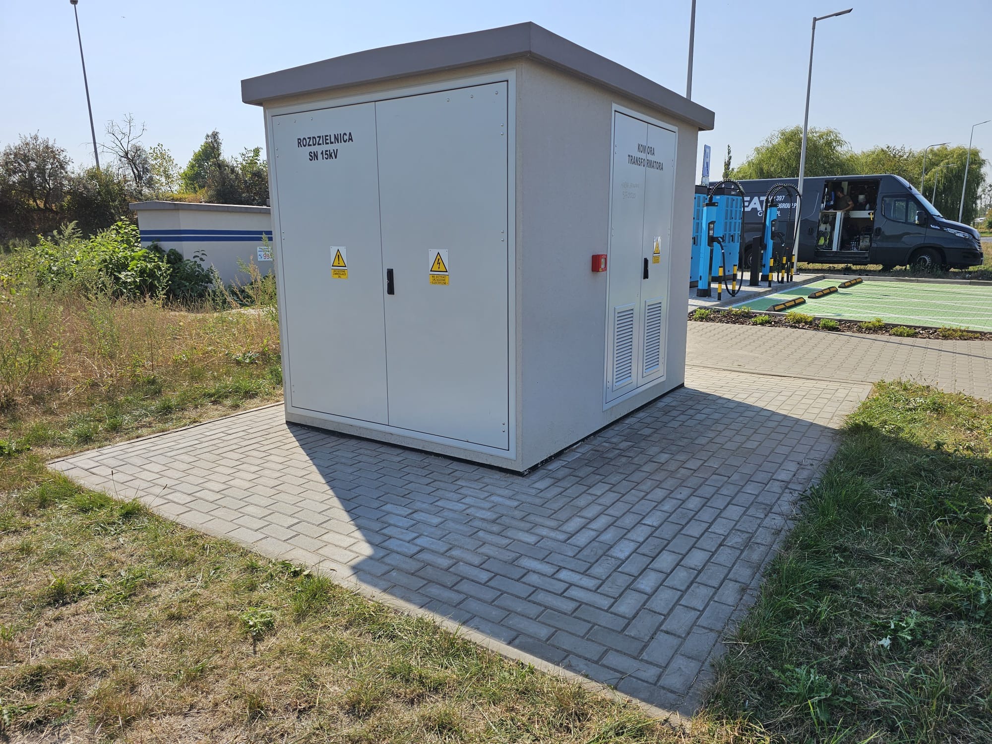 Zewnętrzna stacja transformatorowa SN 15kV z komorą transformatora, posadowiona na kostce brukowej, obok stanowisk ładowania pojazdów elektrycznych i zaparkowanego busa serwisowego.