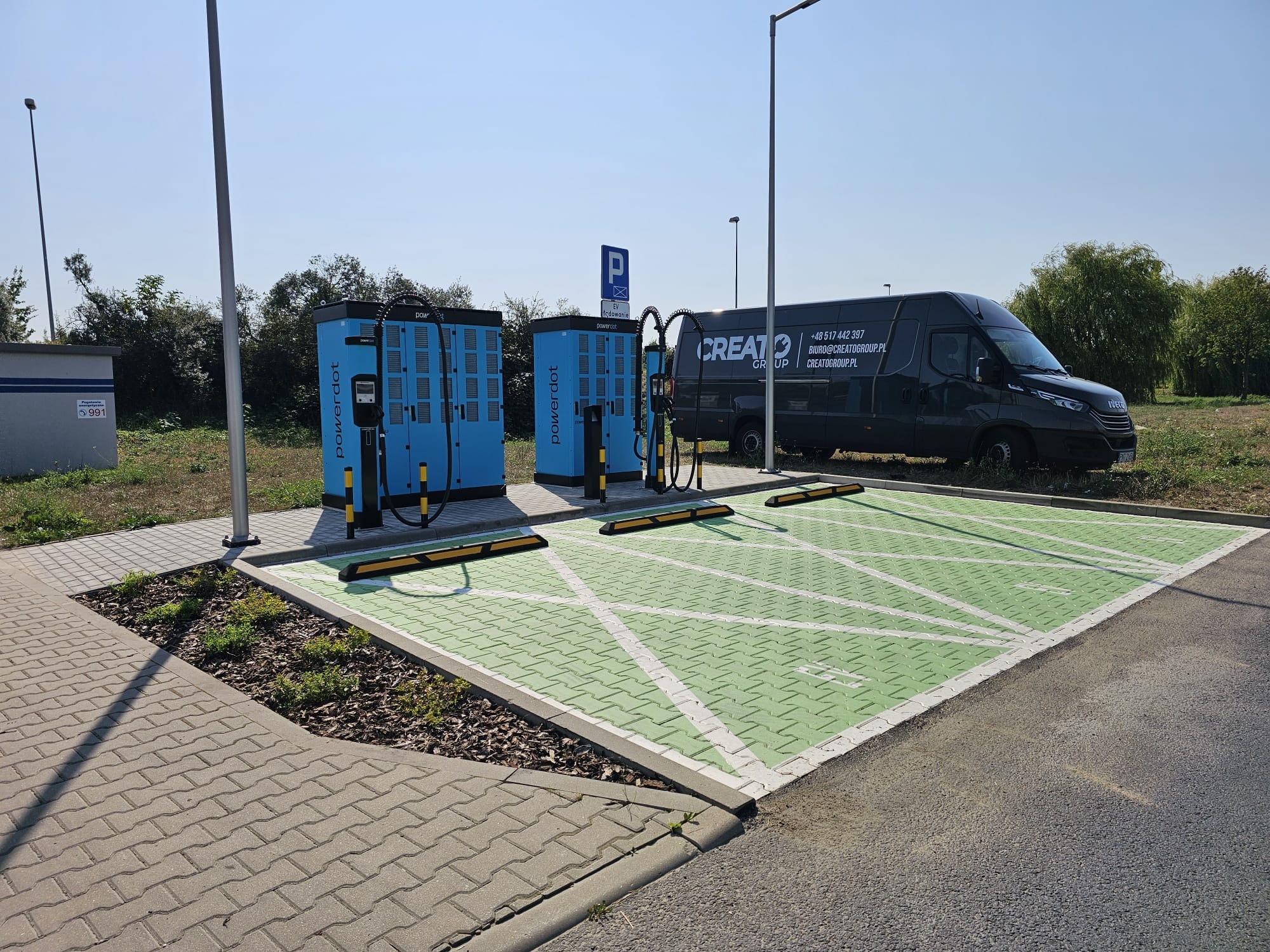 Dwie niebieskie stacje ładowania samochodów elektrycznych Powerdot na zielonym parkingu z białymi oznaczeniami, w tle czarny van firmy Creato Group z numerem telefonu i adresem email.