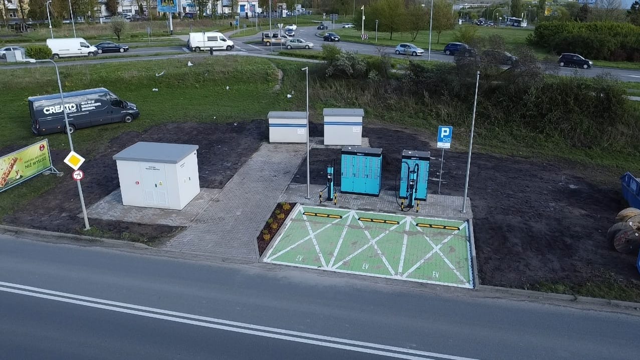 Widok z góry na nowo wybudowaną stację ładowania pojazdów elektrycznych z dwoma punktami ładowania, transformatorem, chodnikiem z kostki brukowej i wyznaczonymi miejscami parkingowymi EV, w tle...