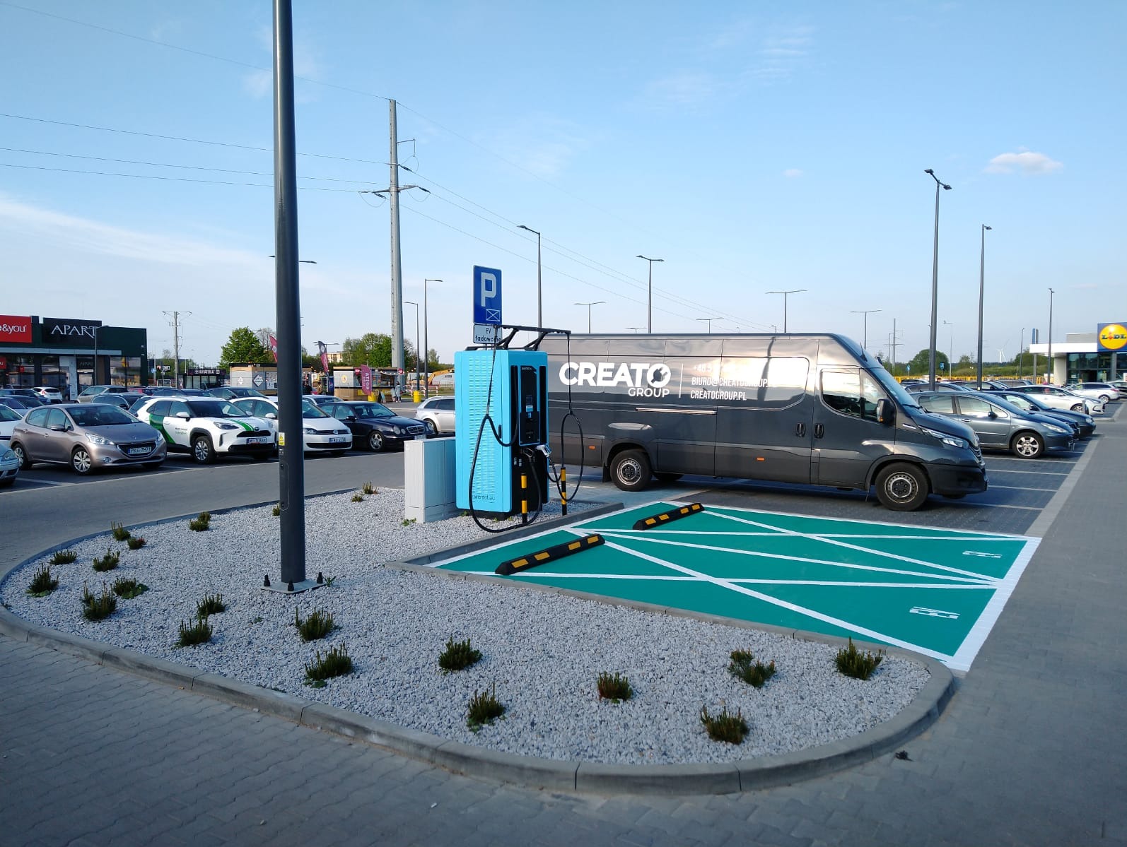 Gotowa stacja ładowania samochodów elektrycznych z niebieską obudową i oznakowanym zielonym miejscem parkingowym, obok zaparkowany szary bus firmy Creato Group na parkingu przed sklepami.