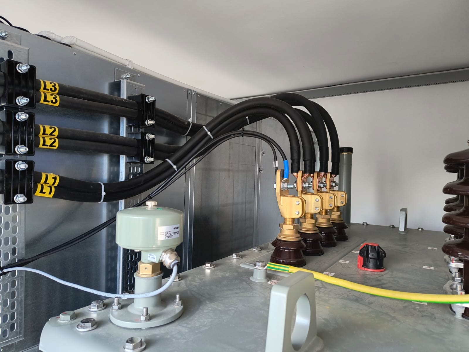 Szczegółowe ujęcie wnętrza transformatora z grubymi kablami oznaczonymi L1, L2, L3, podłączonymi do mosiężnych złączek, widoczne elementy izolacyjne i przewód uziemiający.