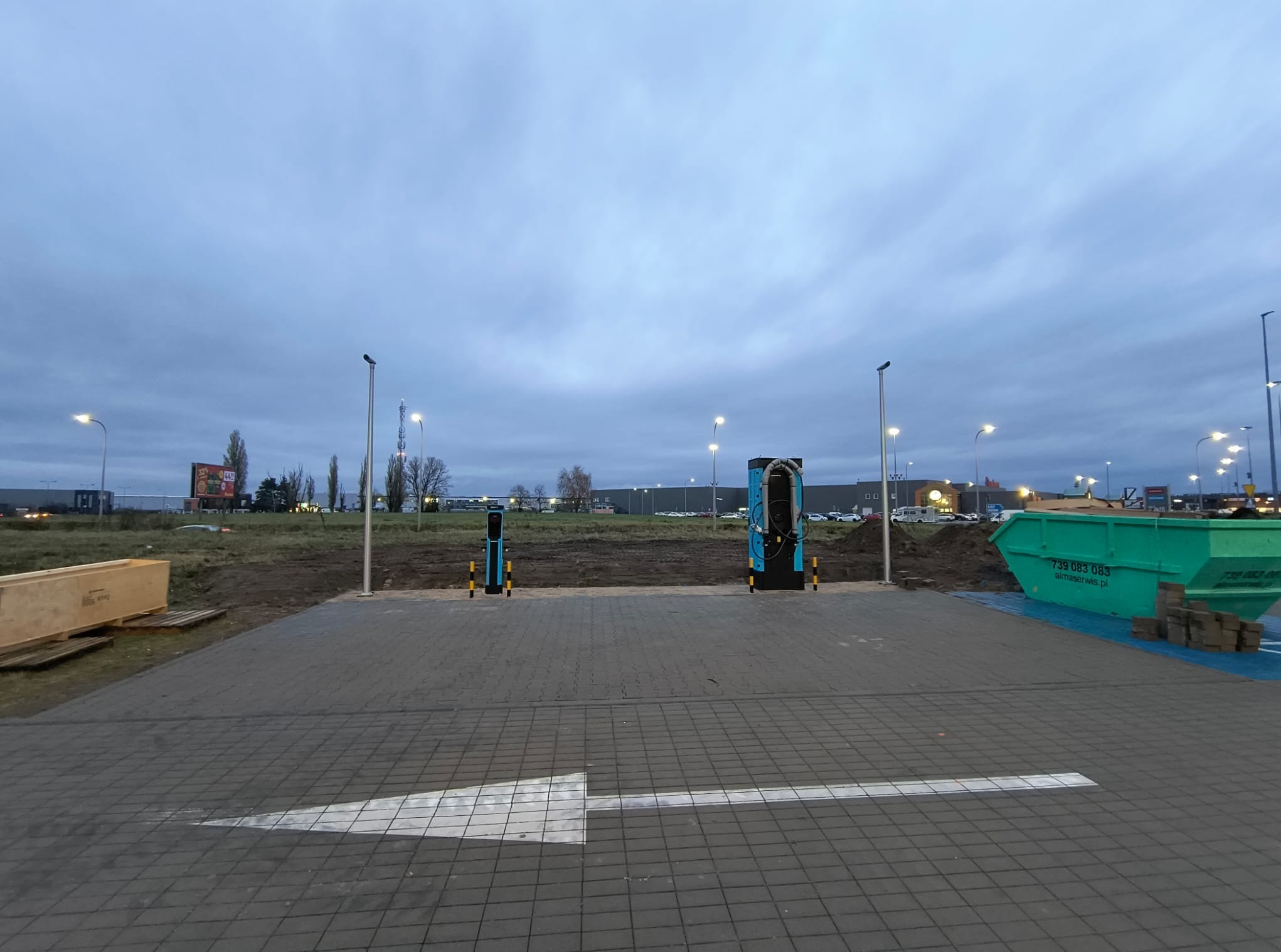 Dwa stanowiska ładowania pojazdów elektrycznych na nowo wybrukowanym placu, strzałka kierunkowa na kostce brukowej, kontener na gruz w tle, pochmurne niebo.