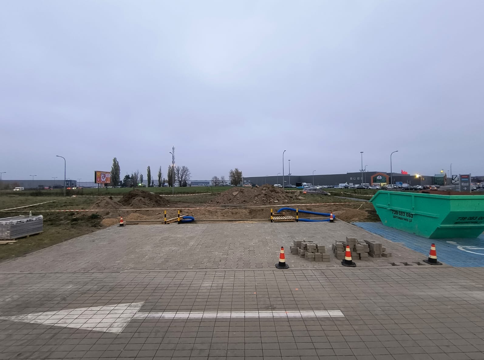 Teren budowy z wykopem, przygotowany podłoże z kostki brukowej, hałdy ziemi, zabezpieczenia, kontener na gruz i budynek w tle.