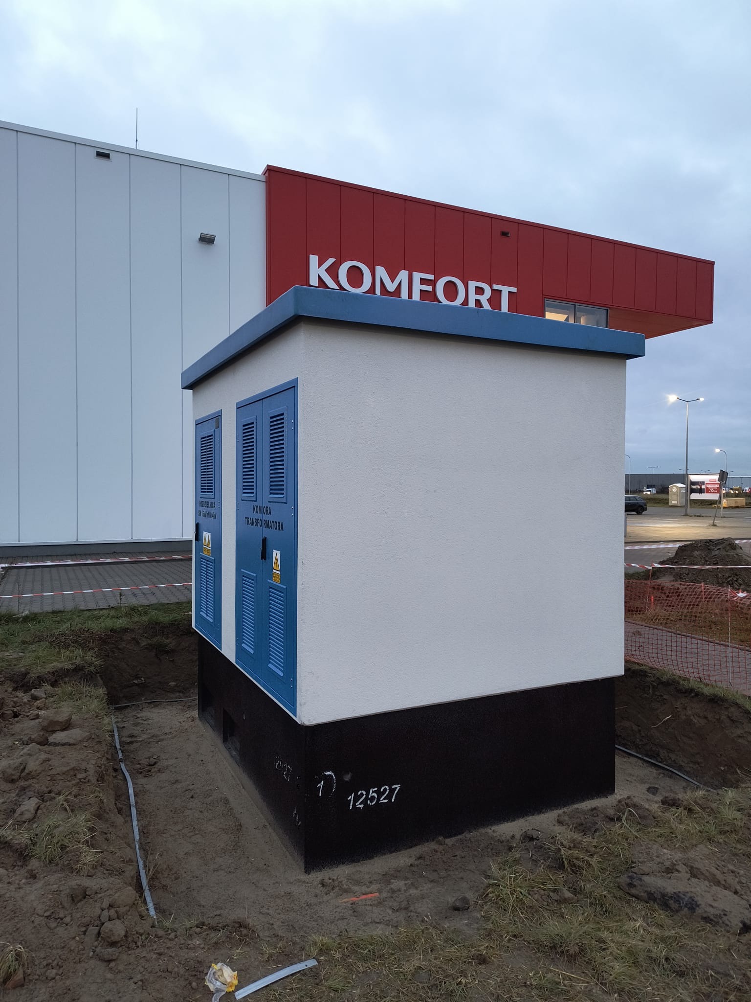 Nowo postawiona, biało-czarna stacja transformatorowa z niebieskimi drzwiami wentylacyjnymi, umieszczona na placu budowy przed budynkiem sklepu Komfort, widoczne kable w ziemi.