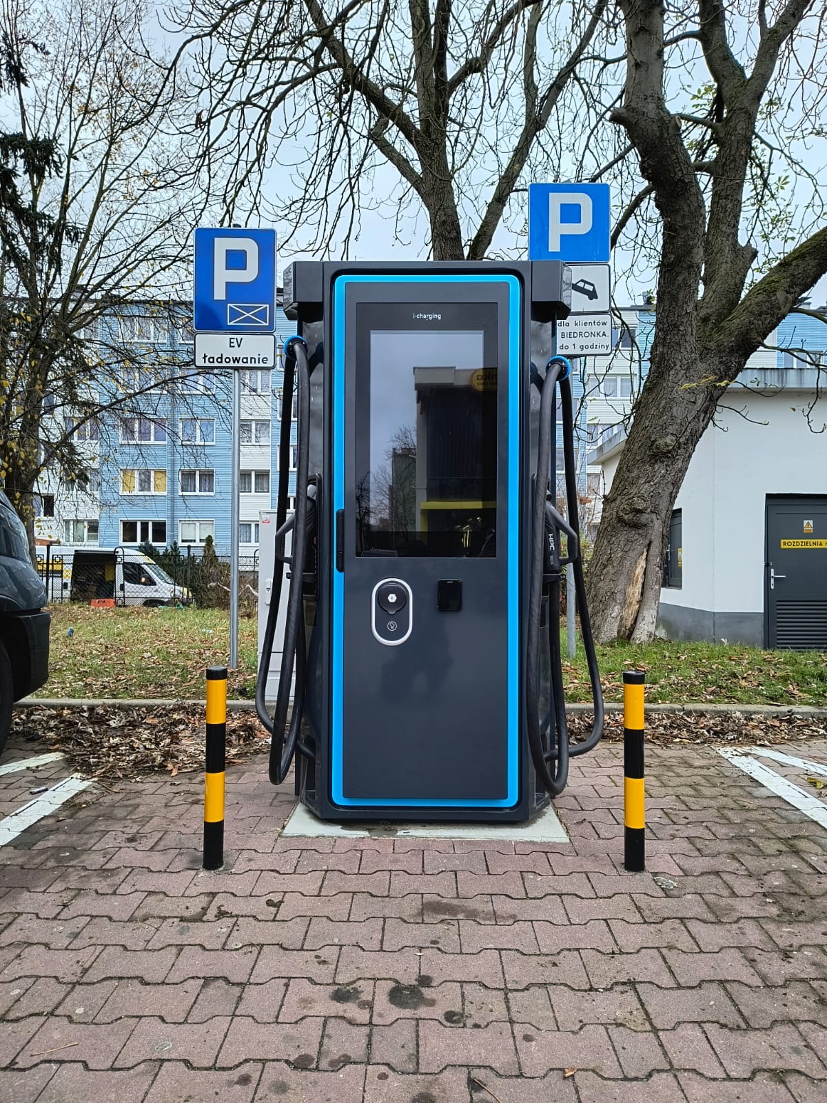 Stacja ładowania pojazdów elektrycznych z niebieską obwódką, ustawiona na brukowanym placu, z widocznymi znakami informacyjnymi i fragmentem drzewa w tle.
