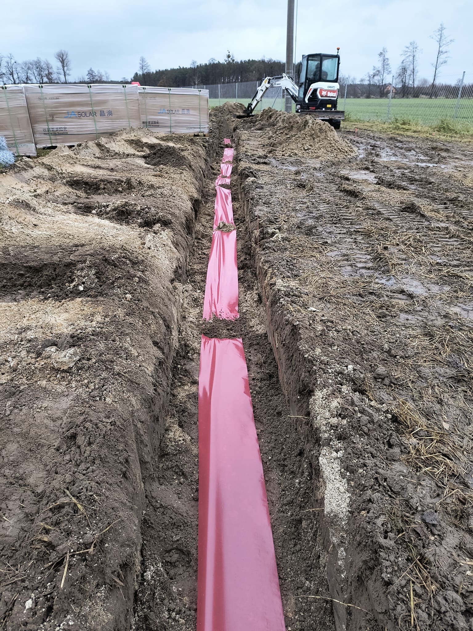 Wykopany rów z ułożonymi czerwonymi rurami ochronnymi, mini koparka Bobcat na tle, palety z panelami solarnymi po lewej stronie, teren błotnisty.