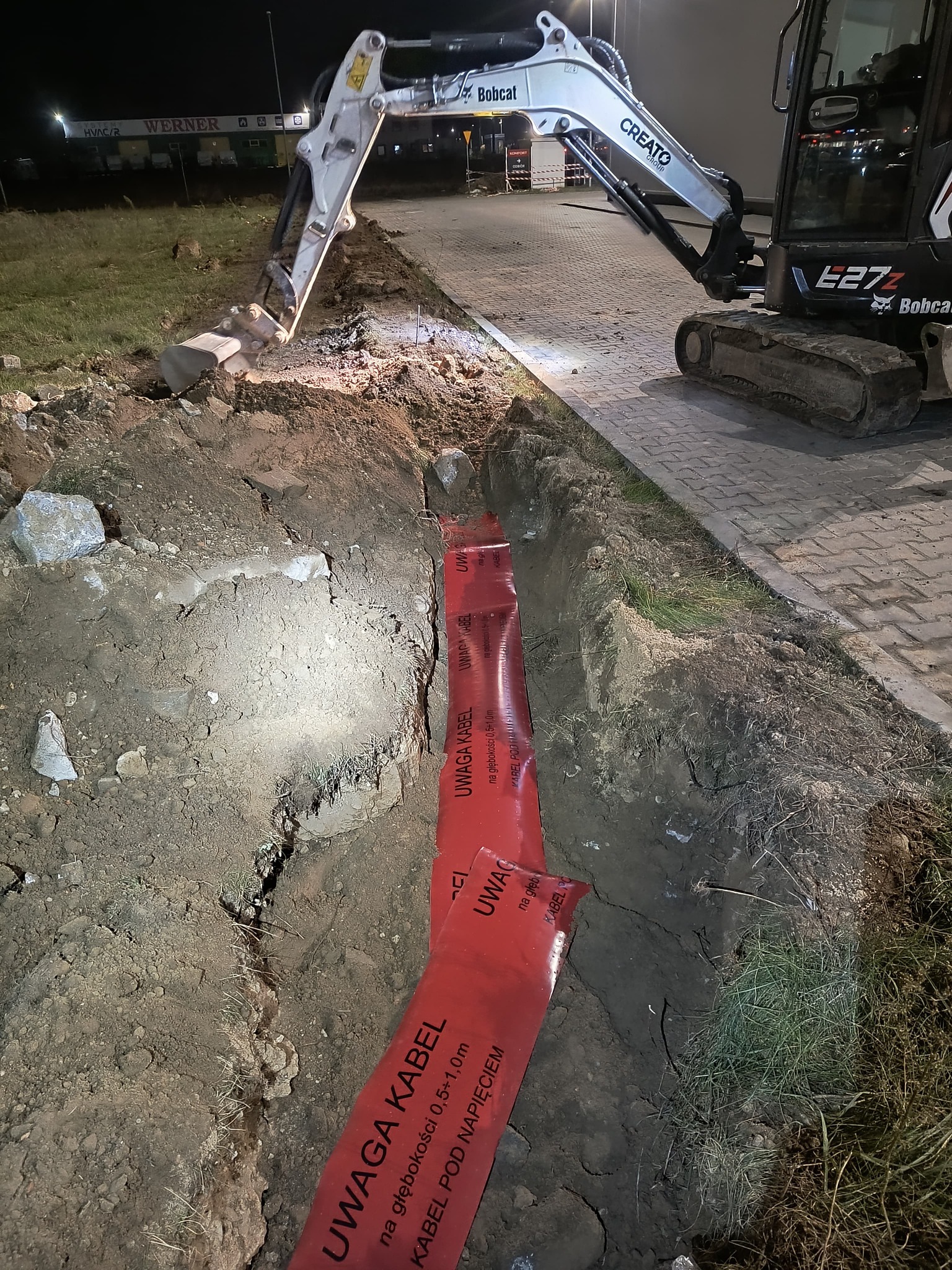 Wykopany rów z ułożoną czerwoną folią ostrzegawczą 'Uwaga Kabel' na głębokości 0,5-1,0m, obok stoi koparka Bobcat E27z, praca wykonywana nocą przy sztucznym oświetleniu.