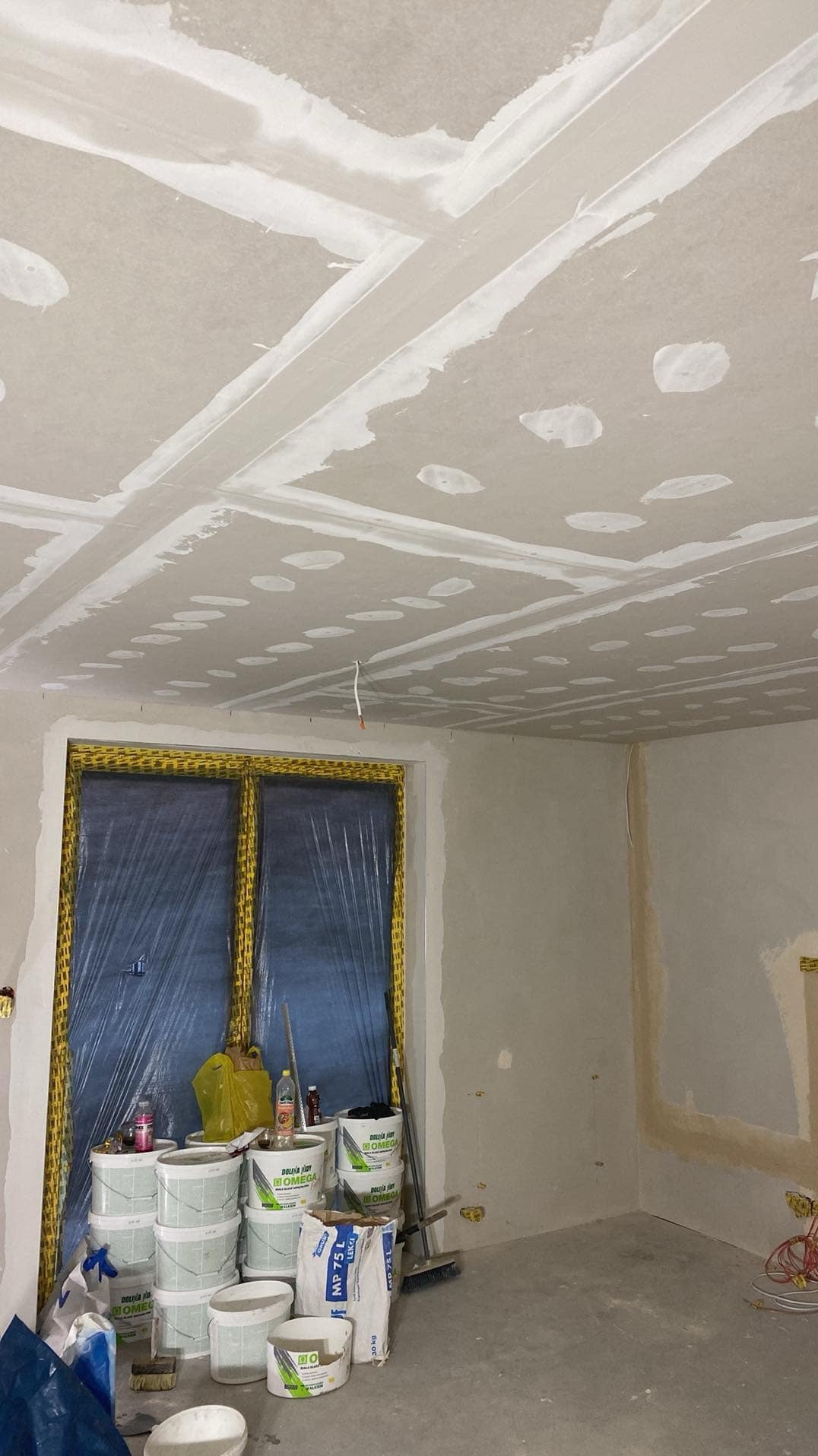 Sufit z płyt gipsowo-kartonowych ze szpachlowanymi łączeniami, widoczne puszki instalacyjne, okno zabezpieczone folią malarską, obok wiadra z masą szpachlową i worki z gipsem