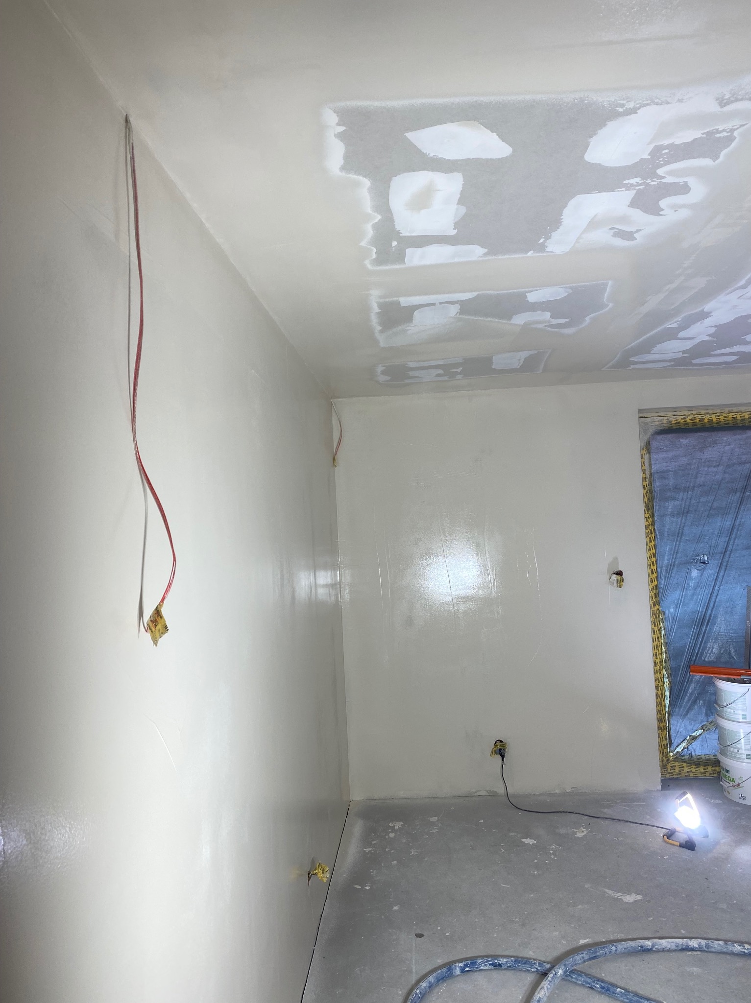 Pokój w trakcie prac wykończeniowych: ściany i sufit po nałożeniu warstwy gładzi, widoczne wystające kable elektryczne zabezpieczone taśmą, podłoga częściowo zakryta, reflektor budowlany oświetla...