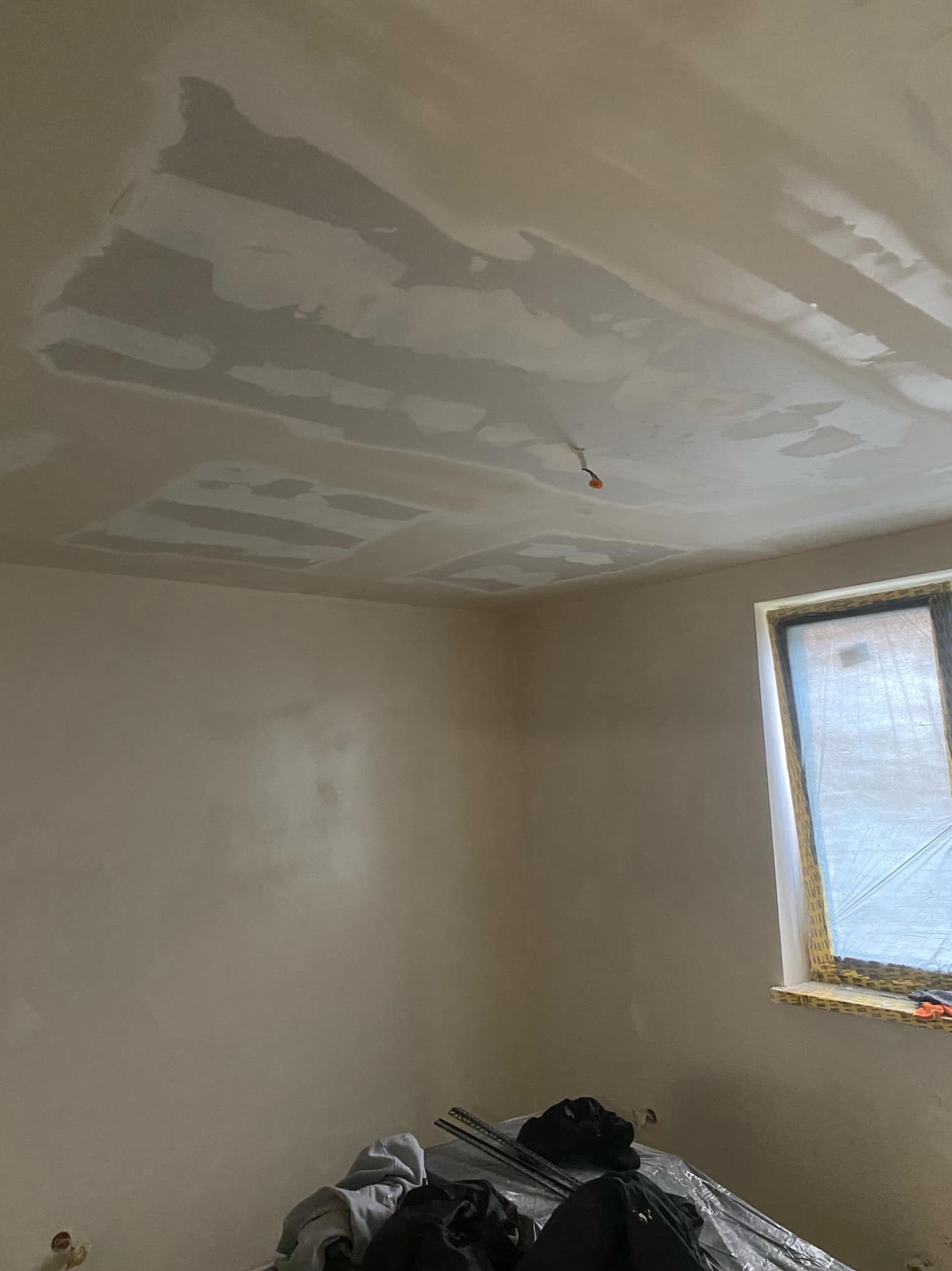 Wnętrze pokoju w trakcie prac wykończeniowych. Na suficie widoczne fragmenty nałożonej i wstępnie wyrównanej warstwy szpachlowej, ściany częściowo pokryte gładzią. Okno zabezpieczone folią malarską...