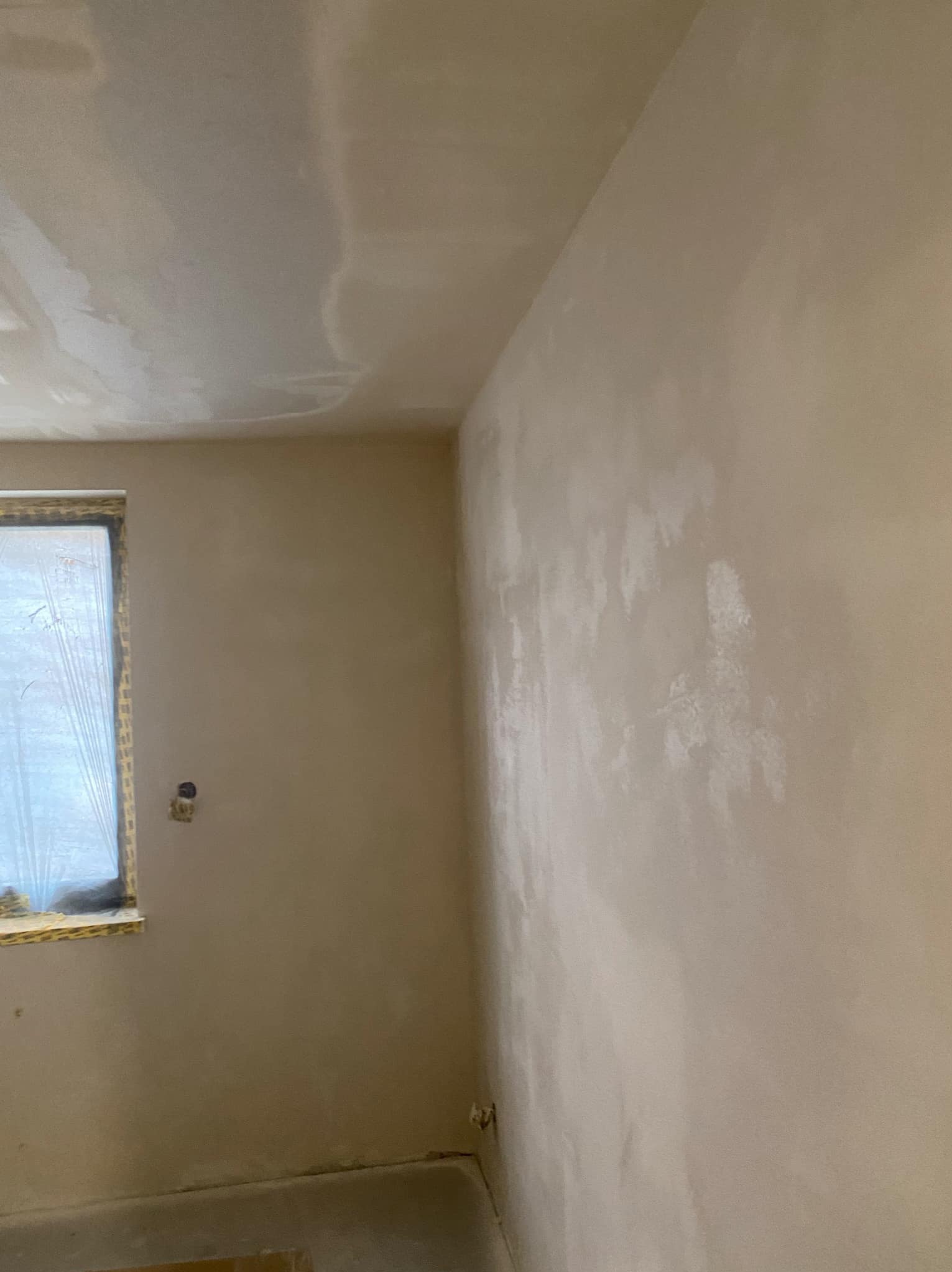Świeżo nałożona, mokra gładź na ścianach i suficie w pokoju, okno zabezpieczone folią malarską, widoczny fragment podłogi.