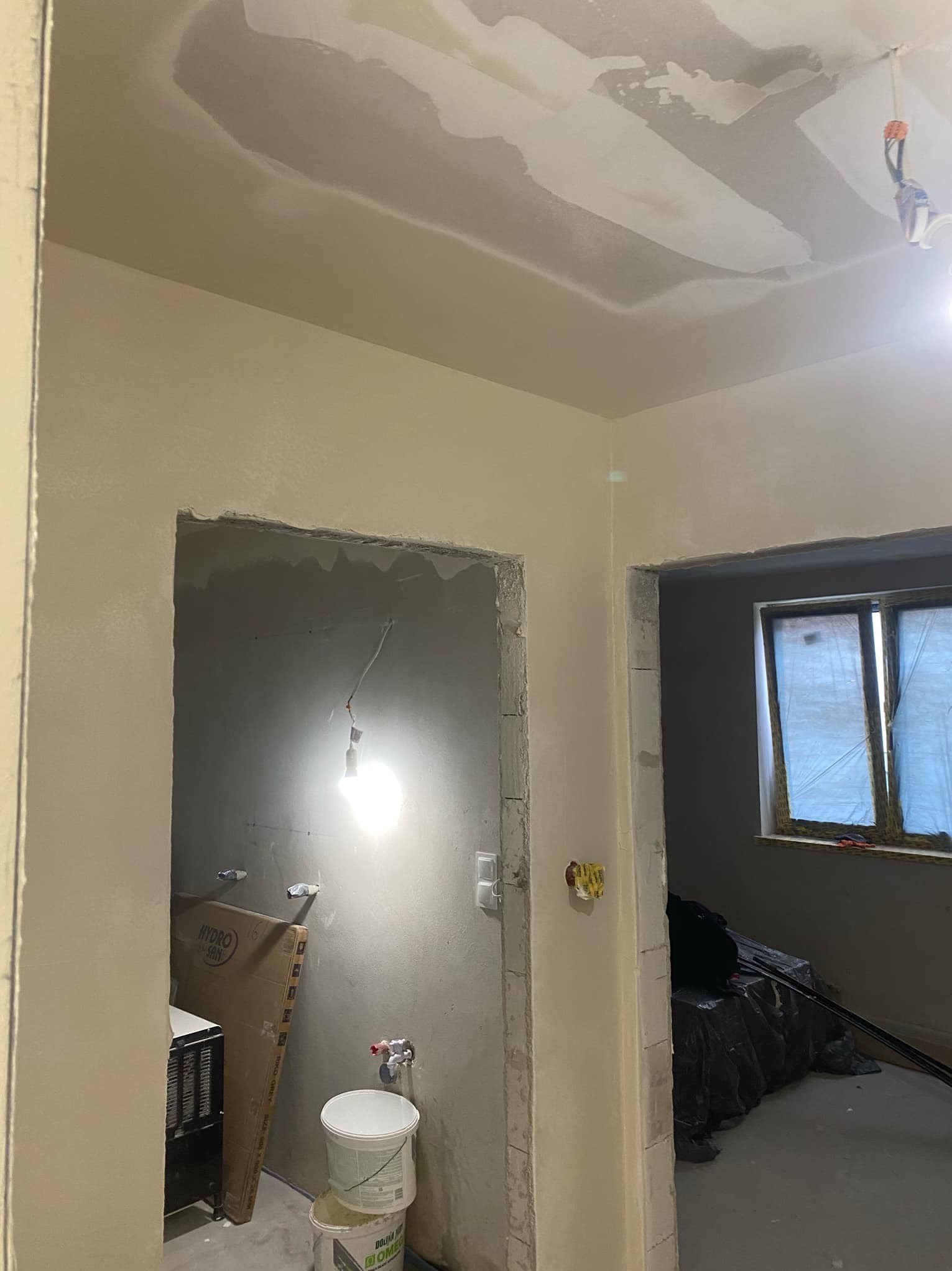 Wnętrze w trakcie prac wykończeniowych: surowy sufit z nałożonymi warstwami zaprawy, przygotowane otwory drzwiowe, ściany częściowo pokryte jasną farbą, widoczne wiadro z zaprawą i kartonowe...