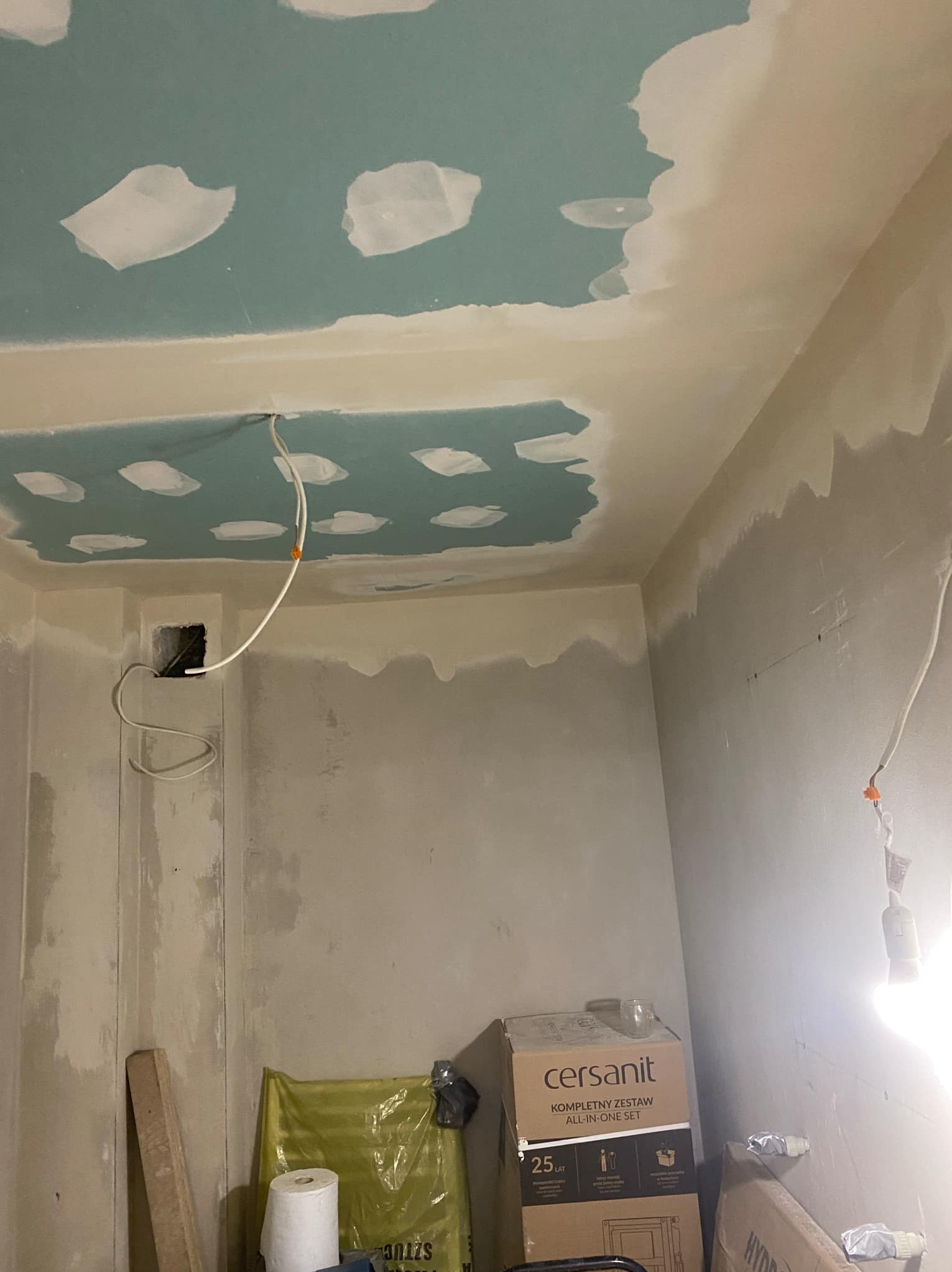 Wnętrze w trakcie prac wykończeniowych: surowy sufit z fragmentami wypełnionymi masą szpachlową i taśmą, ściany przygotowane pod malowanie, widoczne przewody elektryczne i akcesoria budowlane,...