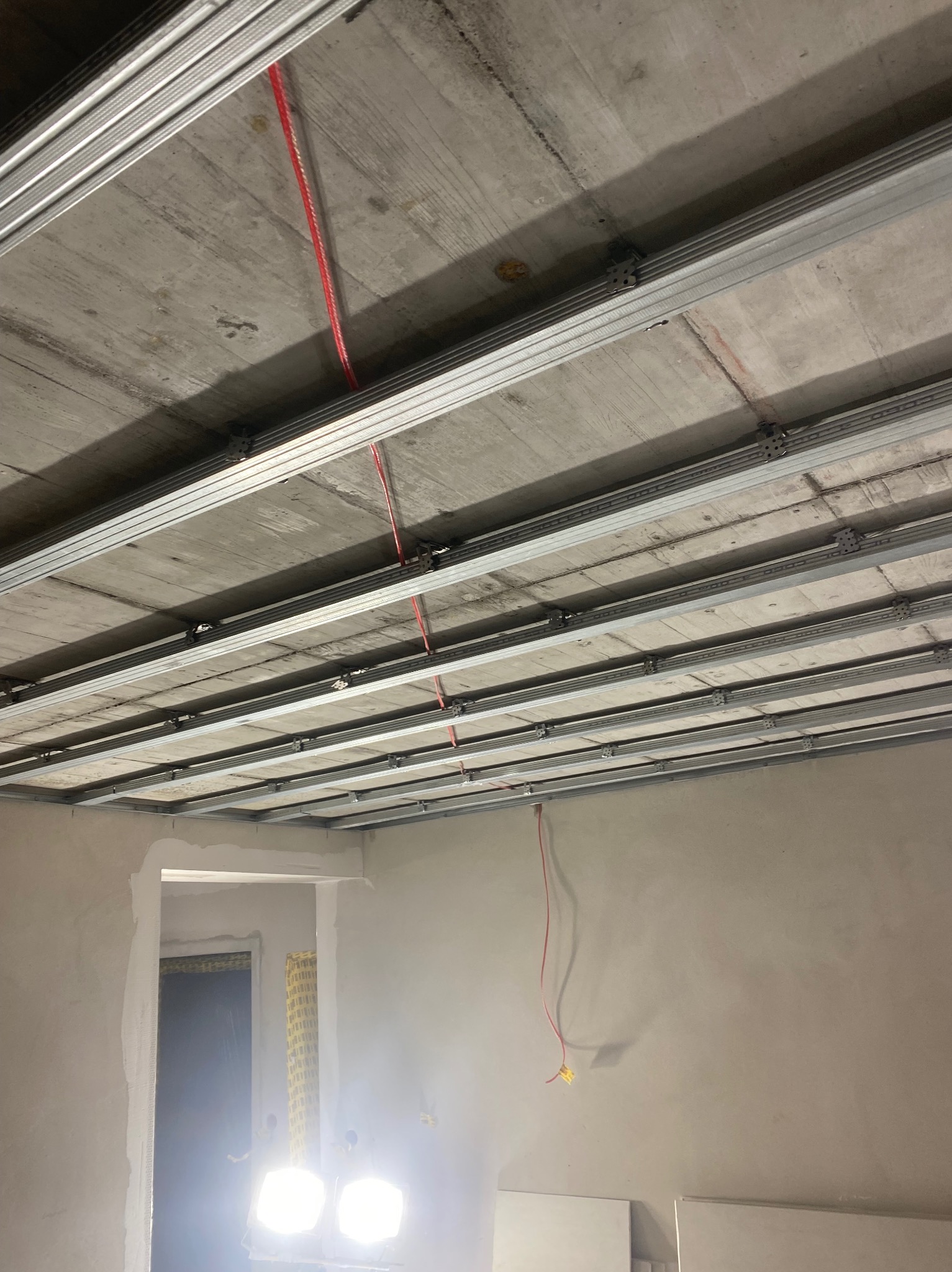 Montaż metalowej konstrukcji sufitu podwieszanego w surowym wnętrzu, widoczne kable elektryczne i przygotowane ściany do dalszych prac wykończeniowych.