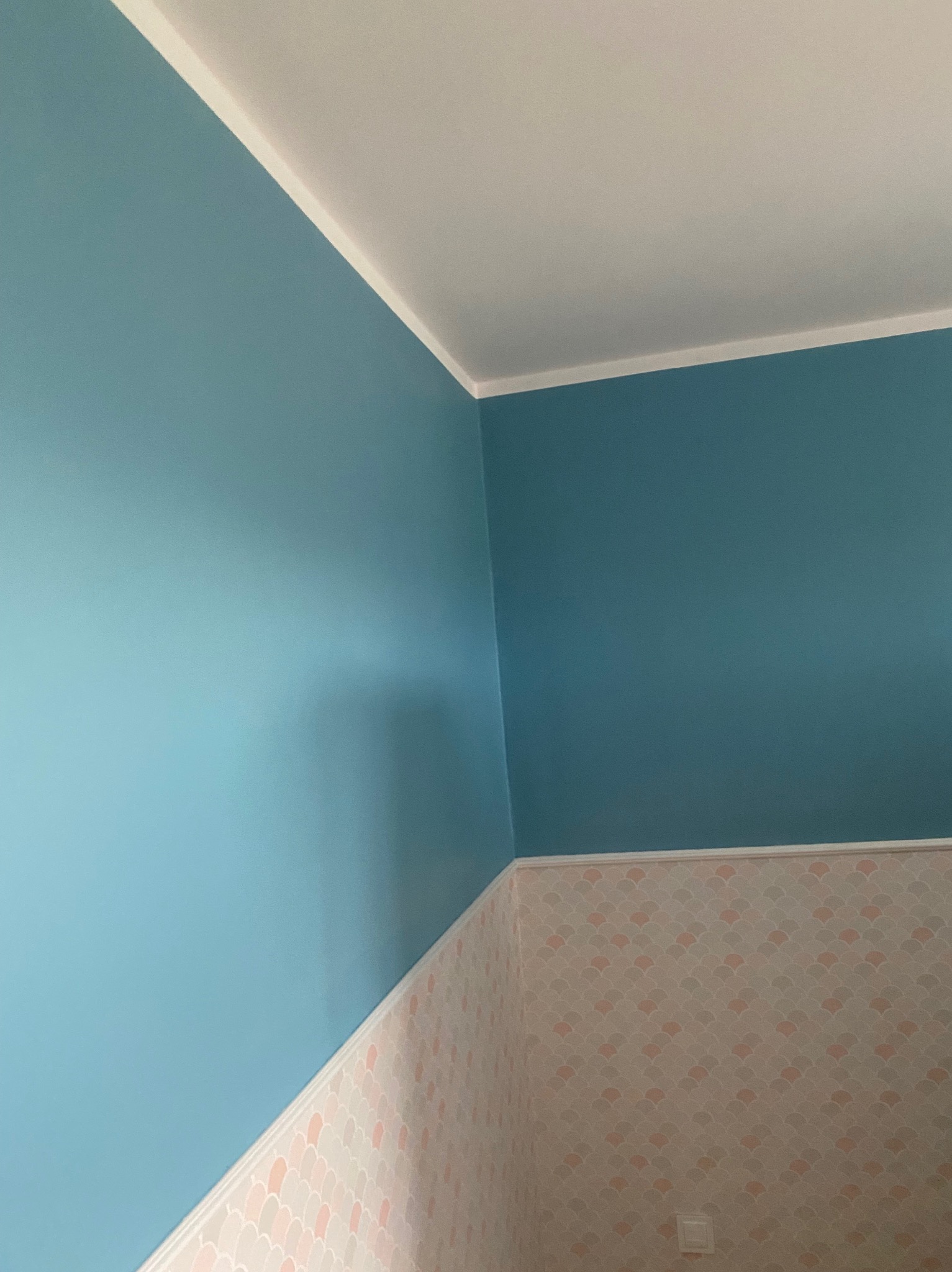 Ściana pomalowana na niebiesko w rogu pomieszczenia, z tapetą w pastelowe wzory na dole i białą listwą przypodłogową oraz sufitową.