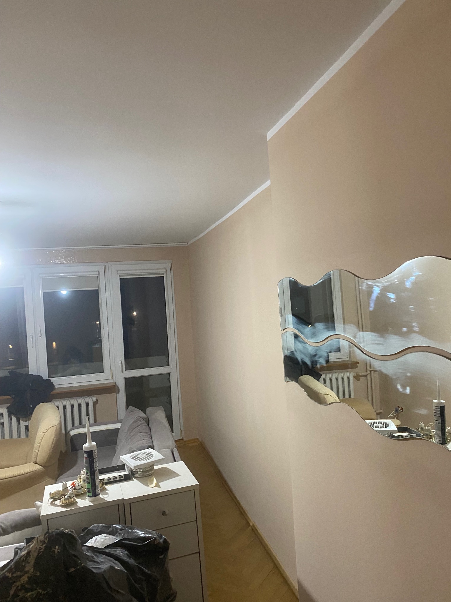 Beżowa ściana po malowaniu z ozdobnym lustrem, widoczne narzędzia i akcesoria malarskie na szafce w tle, okno z grzejnikiem, fragmenty mebli.