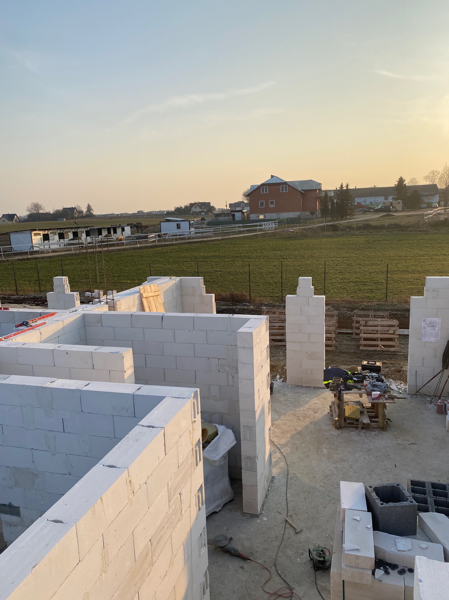 Wznoszone ściany domu z białych bloczków, widok z góry na plac budowy w promieniach zachodzącego słońca, widoczne narzędzia i palety w tle krajobraz wiejski.