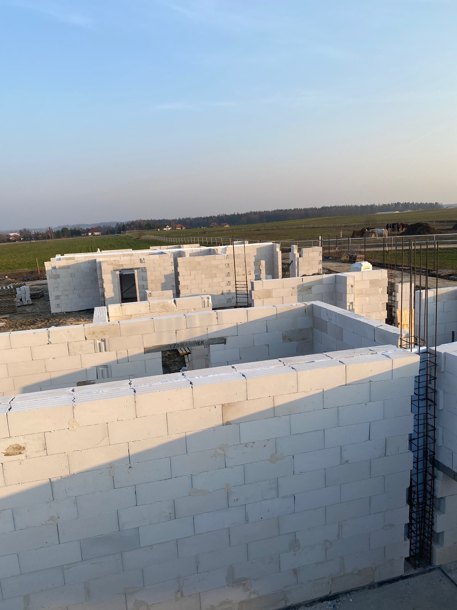Wznoszenie ścian domu z białych bloczków na tle zielonego pola i błękitnego nieba, widoczne zbrojenie pionowe.
