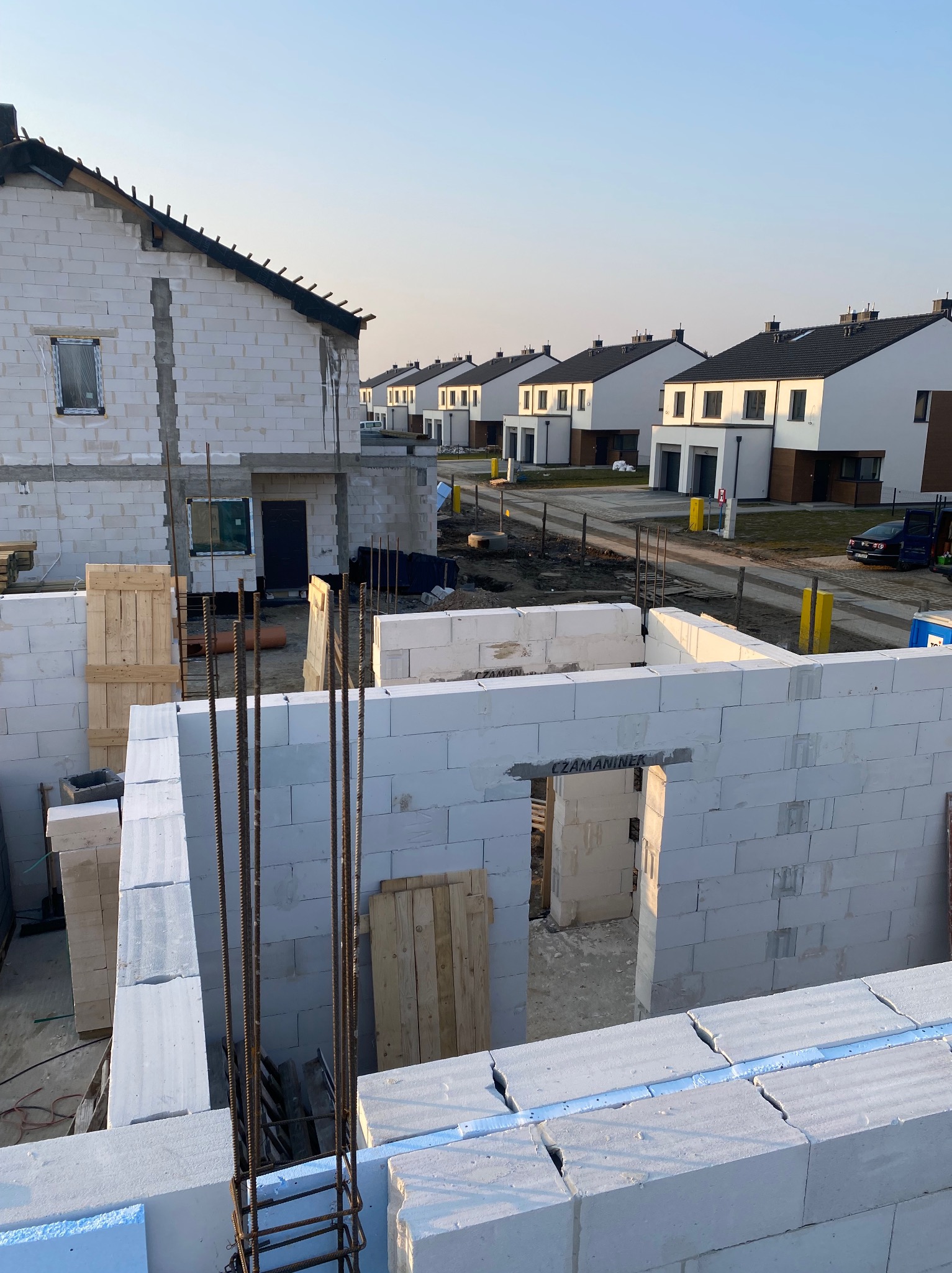 Wznoszenie ścian parterowego domu z białych bloczków, widoczne zbrojenie i szpaler identycznych domów w tle, wczesny etap budowy osiedla.