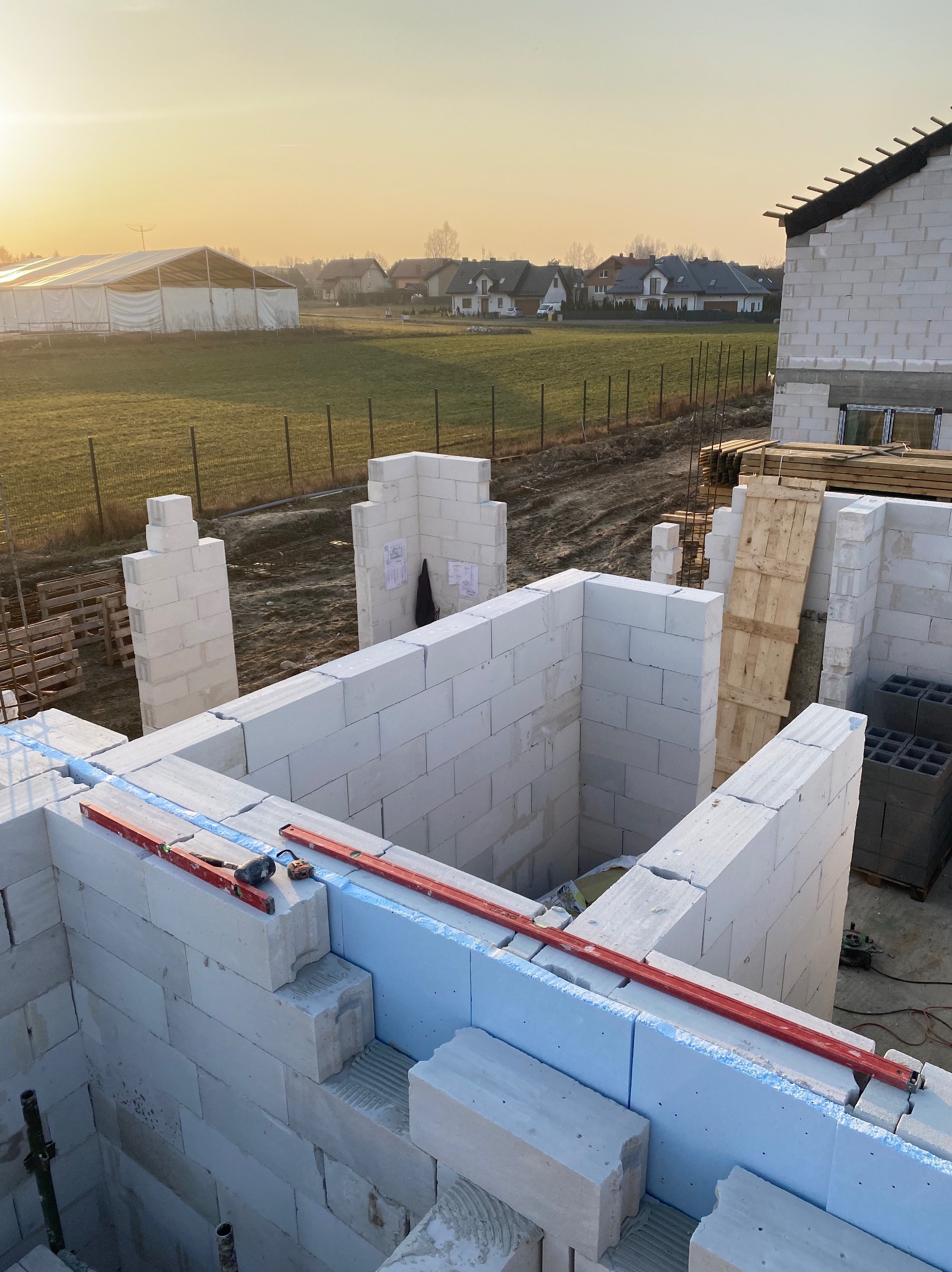 Wznoszenie ścian zewnętrznych domu z betonu komórkowego, widoczne narzędzia murarskie, poziomica, ocieplenie pionowe styropianem, w tle zabudowa jednorodzinna i hala namiotowa oświetlone...