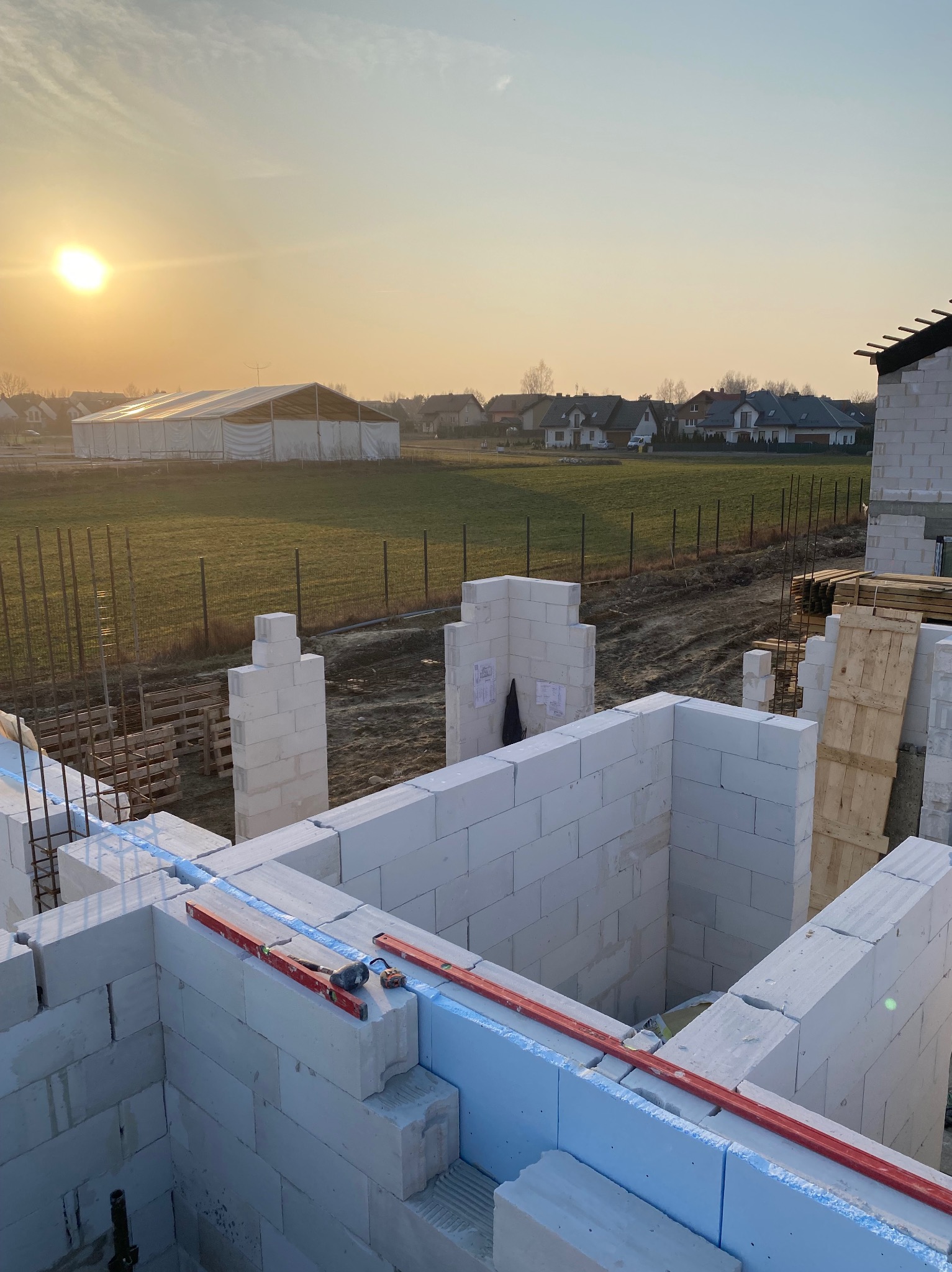 Wznoszenie ścian domu z bloczków, widoczne poziomice, zaprawa klejowa, zbrojenia, w tle zabudowa jednorodzinna i zachodzące słońce.