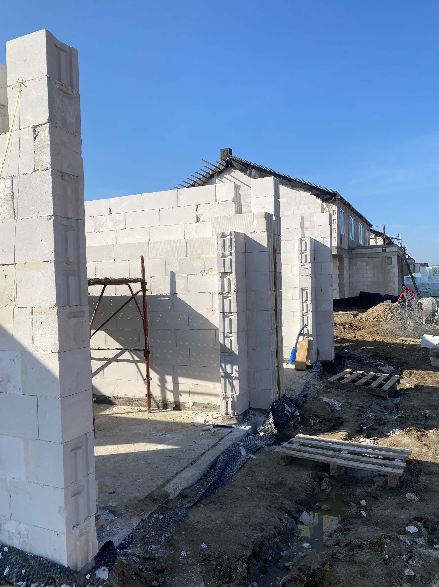 Wznoszenie ścian domu z białych bloczków w Kartuzach, widoczne rusztowanie i przygotowane palety, etap budowy w słoneczny dzień.