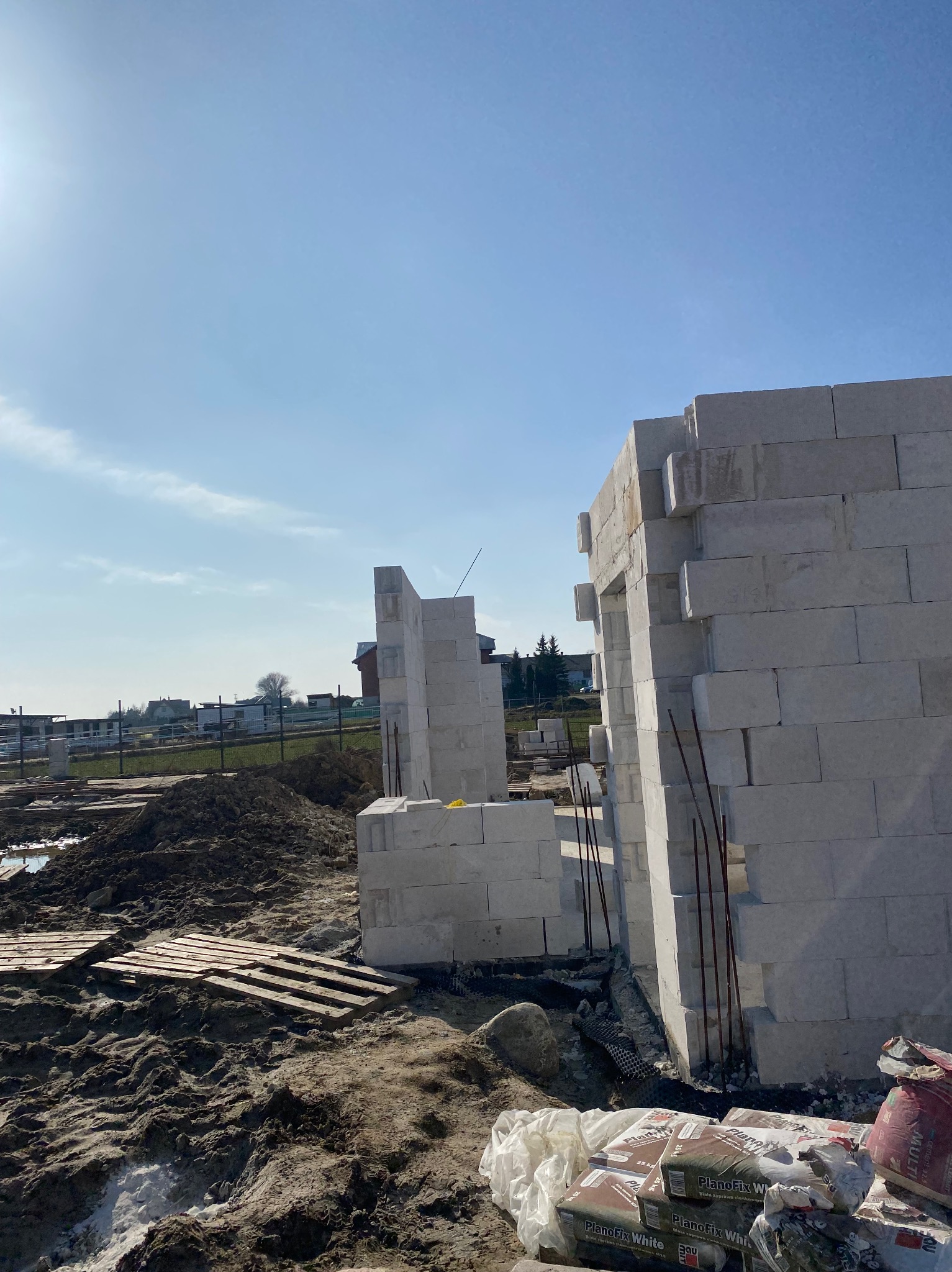 Wznoszenie ścian domu z białych bloczków betonu komórkowego, widoczne pręty zbrojeniowe, palety na błotnistym terenie oraz worki z zaprawą murarską PlanoFix White.